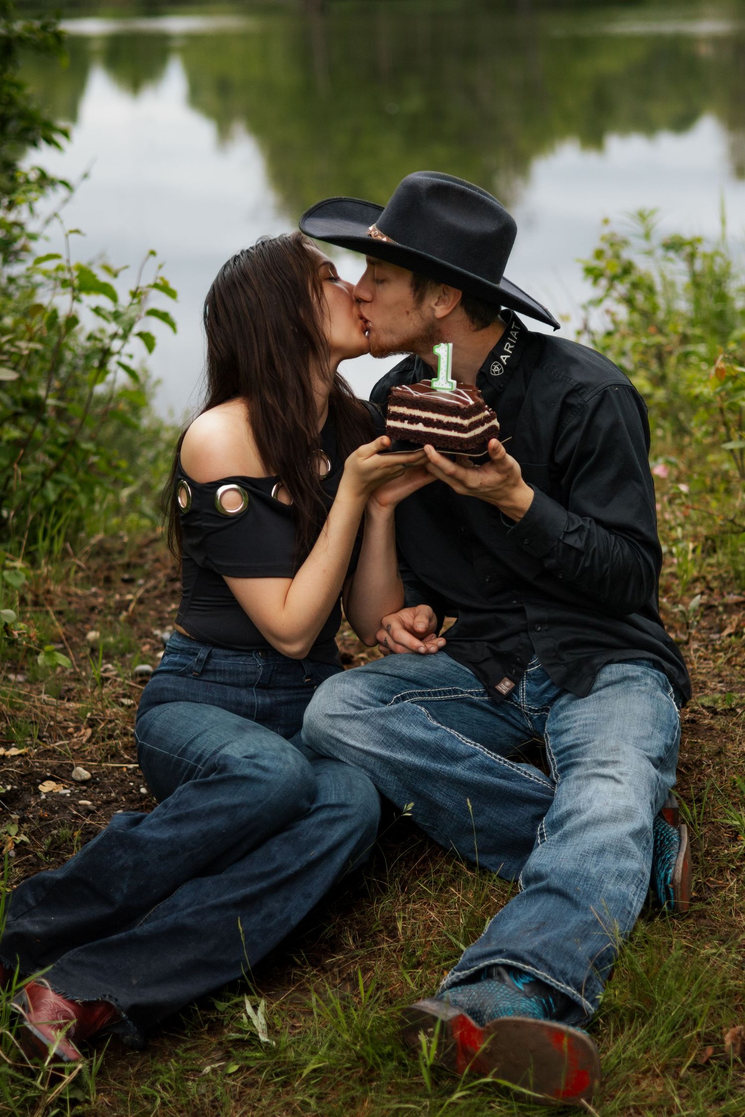Couple kissing outdoors by a lake, sharing a birthday cake with candle, wearing cowboy hats and jeans.