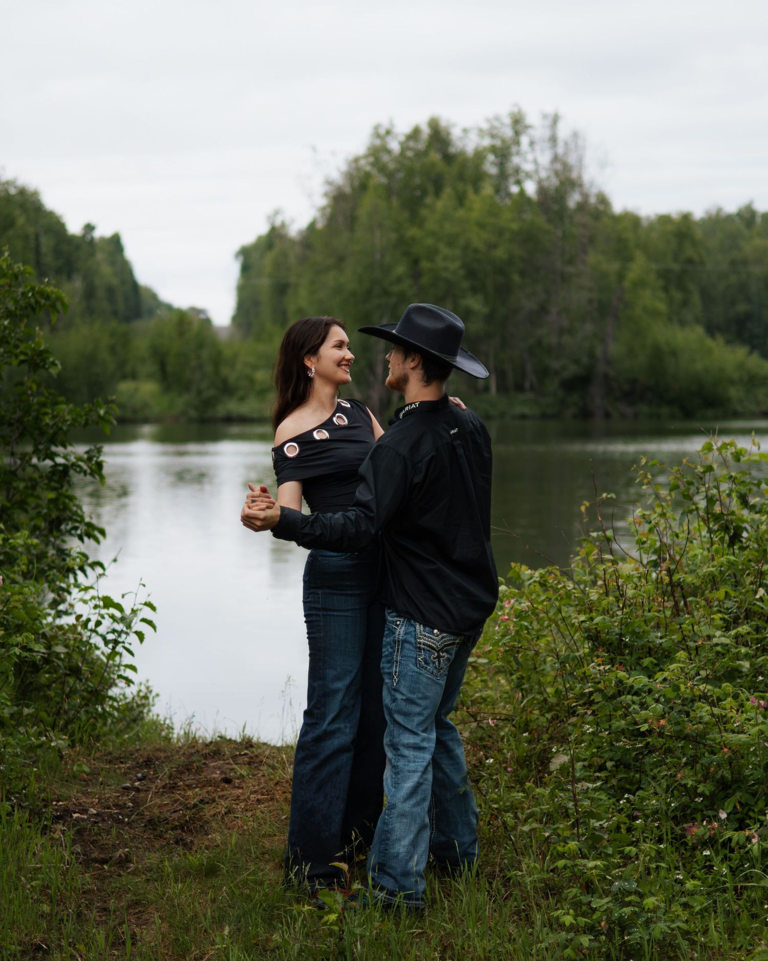 Couple dancing near a serene lake surrounded by lush green trees on an overcast day.