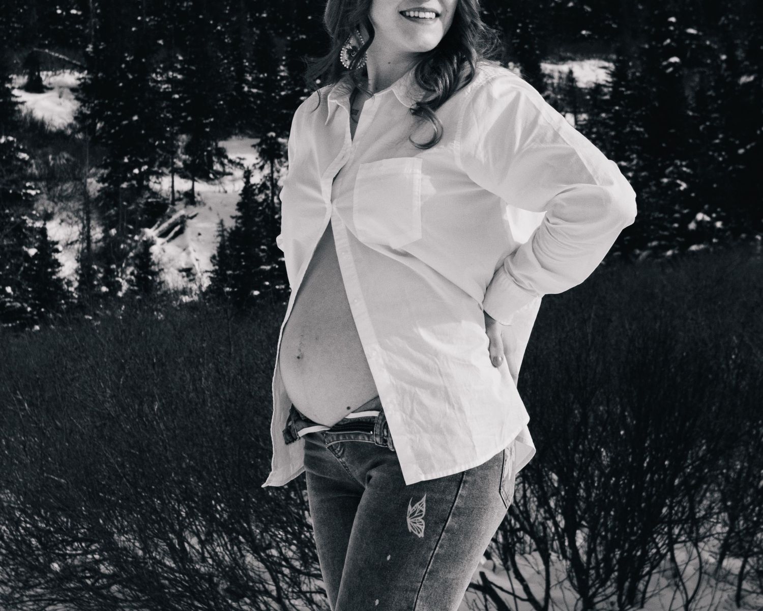 Black and white maternity photo of smiling pregnant woman in white shirt and jeans outdoors in winter snow.