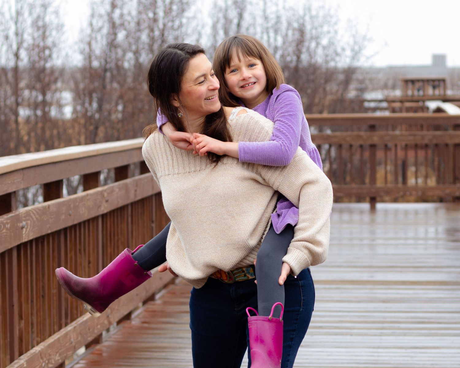 A woman gives a smiling girl a piggyback ride on a wooden bridge, both wearing pink rain boots.