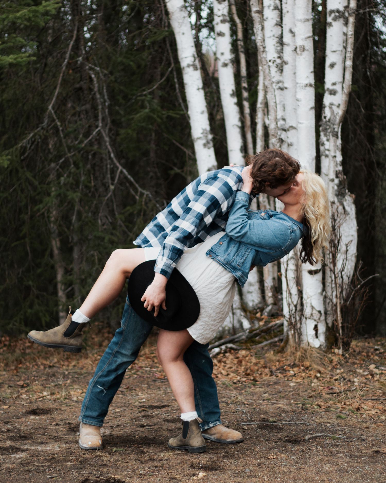 Couple sharing a romantic dip kiss among birch trees in a forest, wearing casual denim and white outfits.