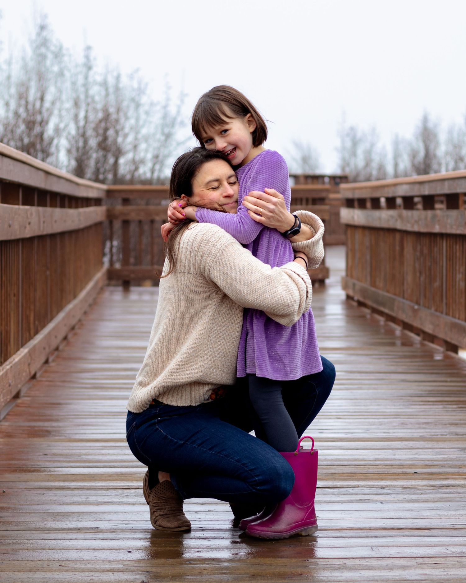 A mother kneels on a wooden bridge hugging her smiling young daughter in a purple shirt and pink rain boots.