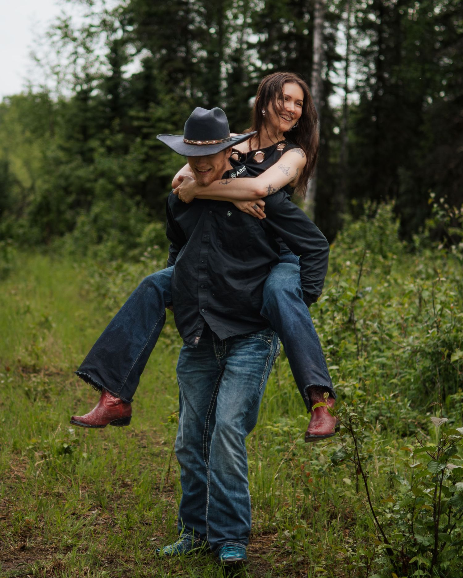 A man in a cowboy hat gives a smiling woman a piggyback ride in a lush green forest setting.