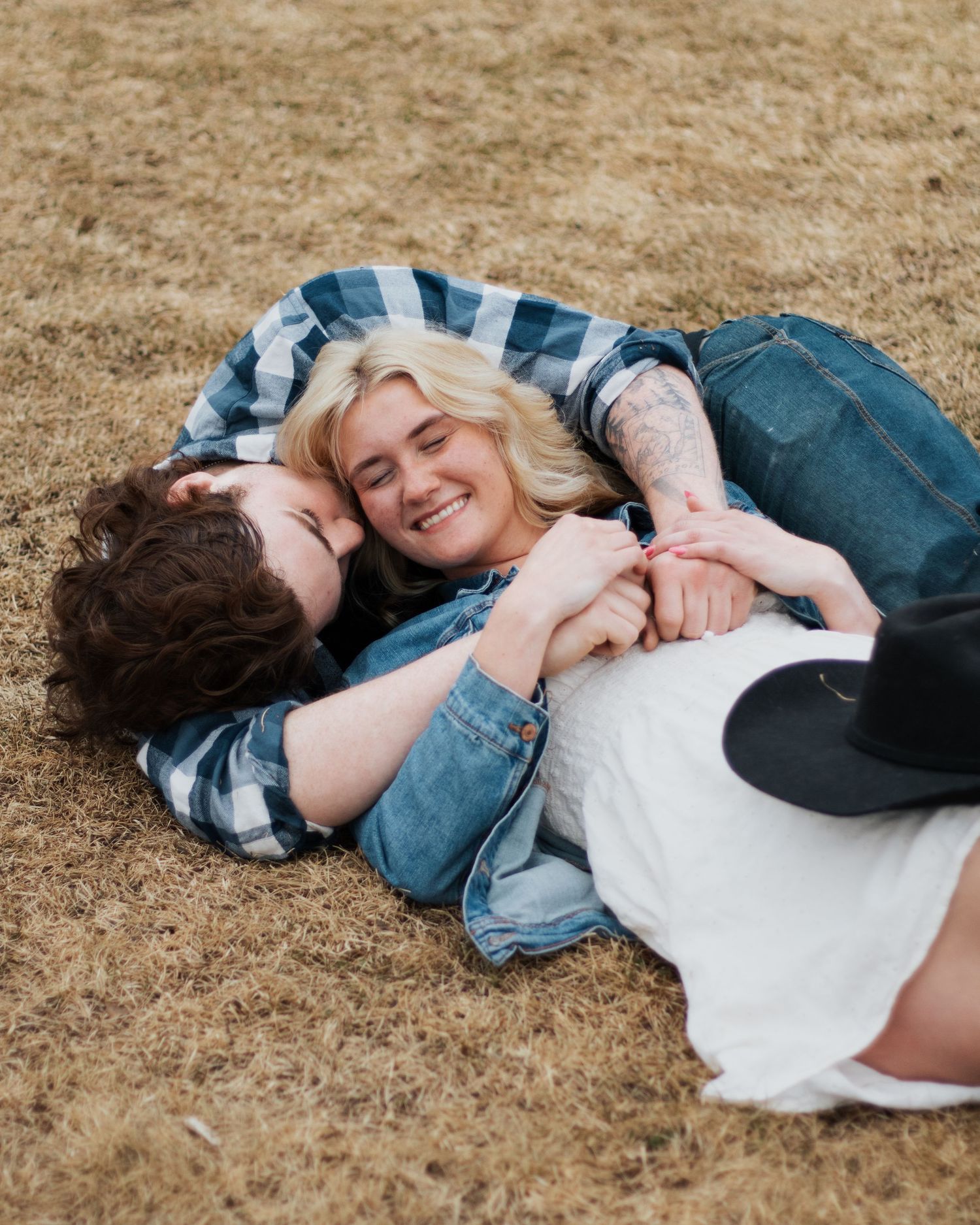 Couple lying on dry grass, smiling and holding hands in a romantic outdoor moment.