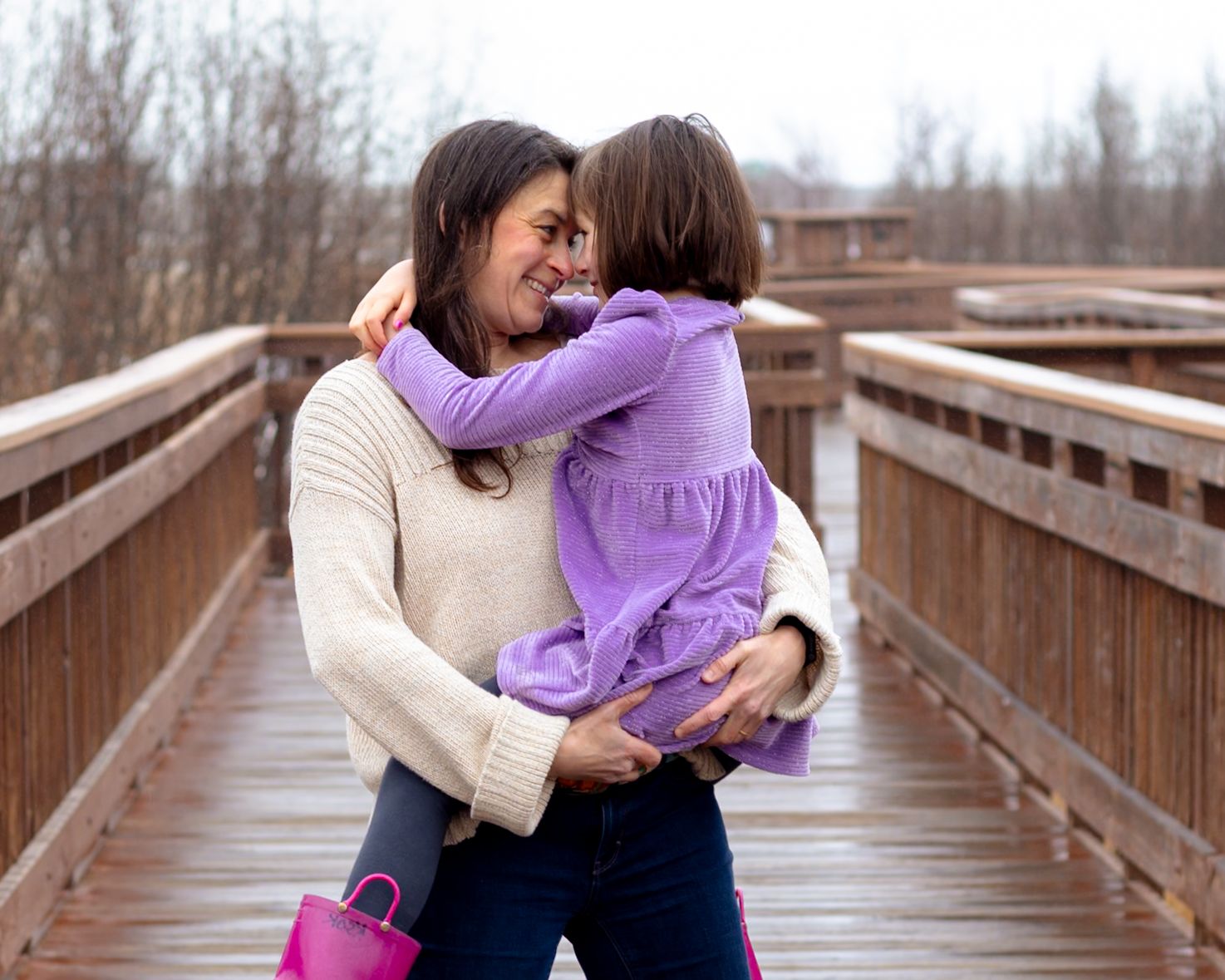 A mother hugs her young daughter on a wooden bridge, both smiling warmly in casual purple and white outfits.