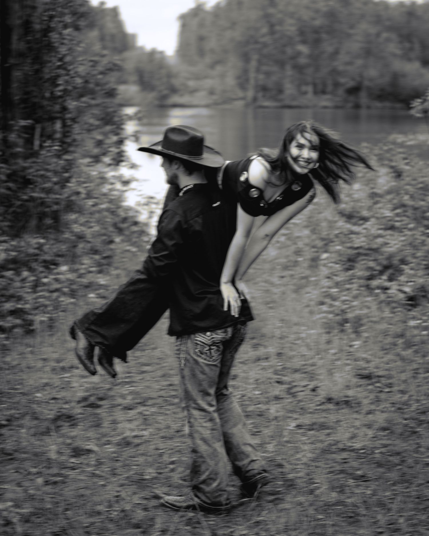 Man in cowboy hat carrying laughing woman on his back near a river in black and white photo.