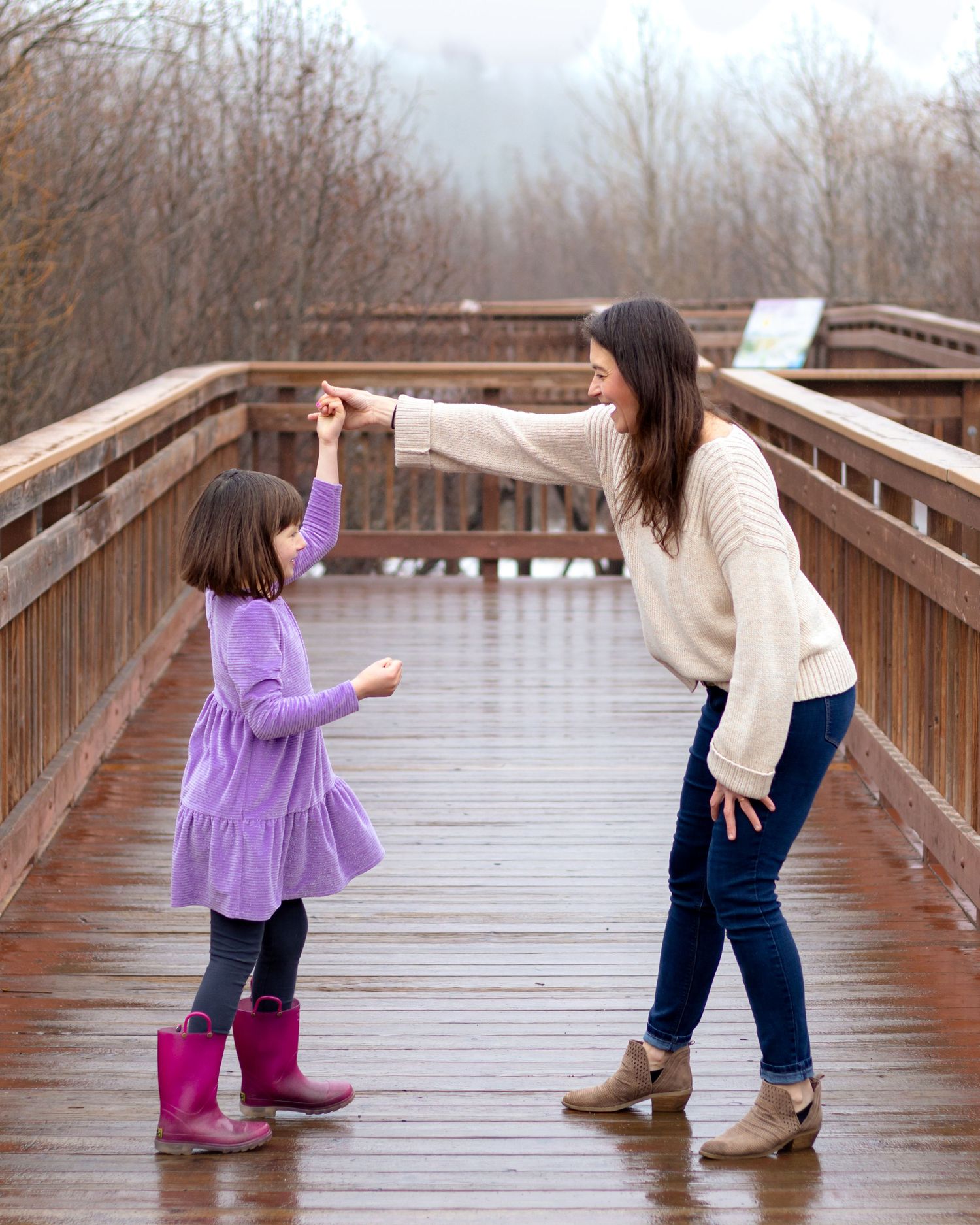 A woman and young girl in purple dress and pink boots dance together on a wooden bridge outdoors.