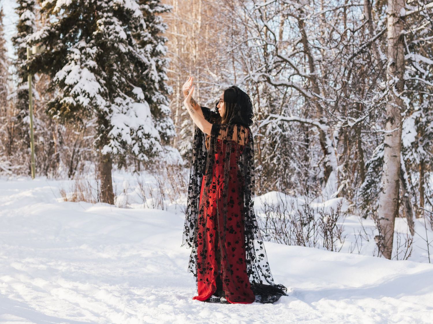 Woman in red and black lace gown poses in snowy winter forest with snow-covered evergreen trees.