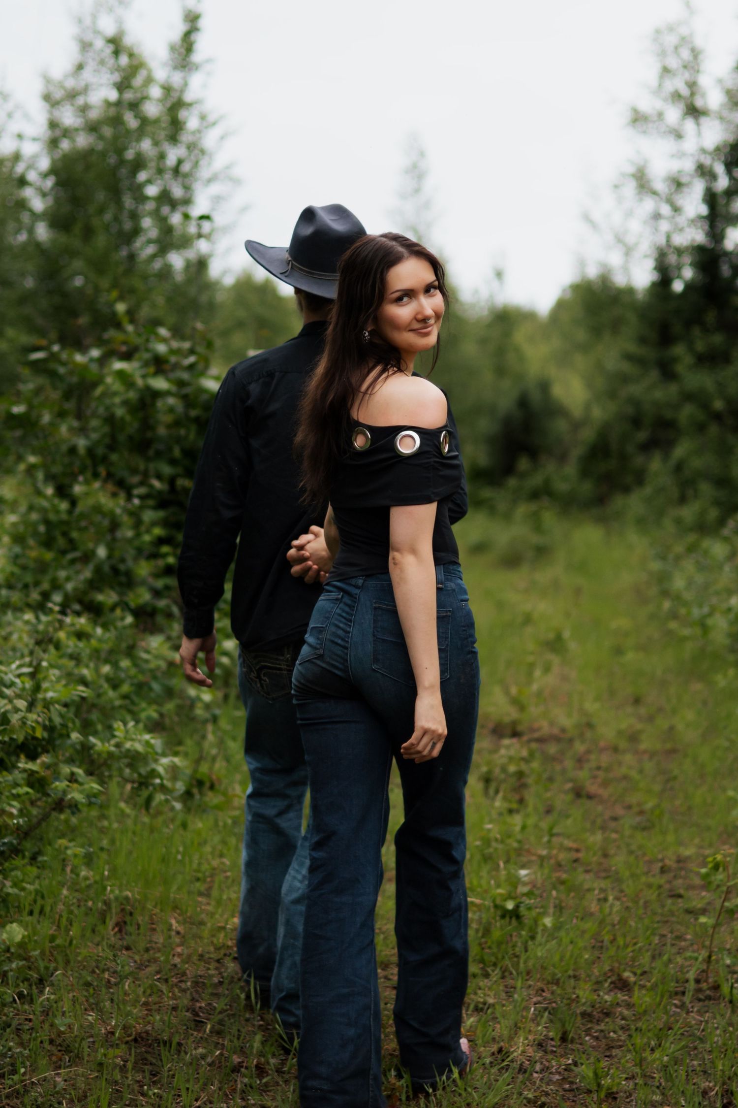 A couple walks hand-in-hand through a lush green path, woman in off-shoulder top looks back smiling, man in black cowboy hat.