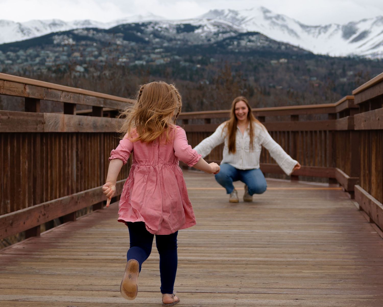 A mother crouches on a wooden bridge with arms open as her young daughter in a pink dress runs toward her with snowy mountains behind.