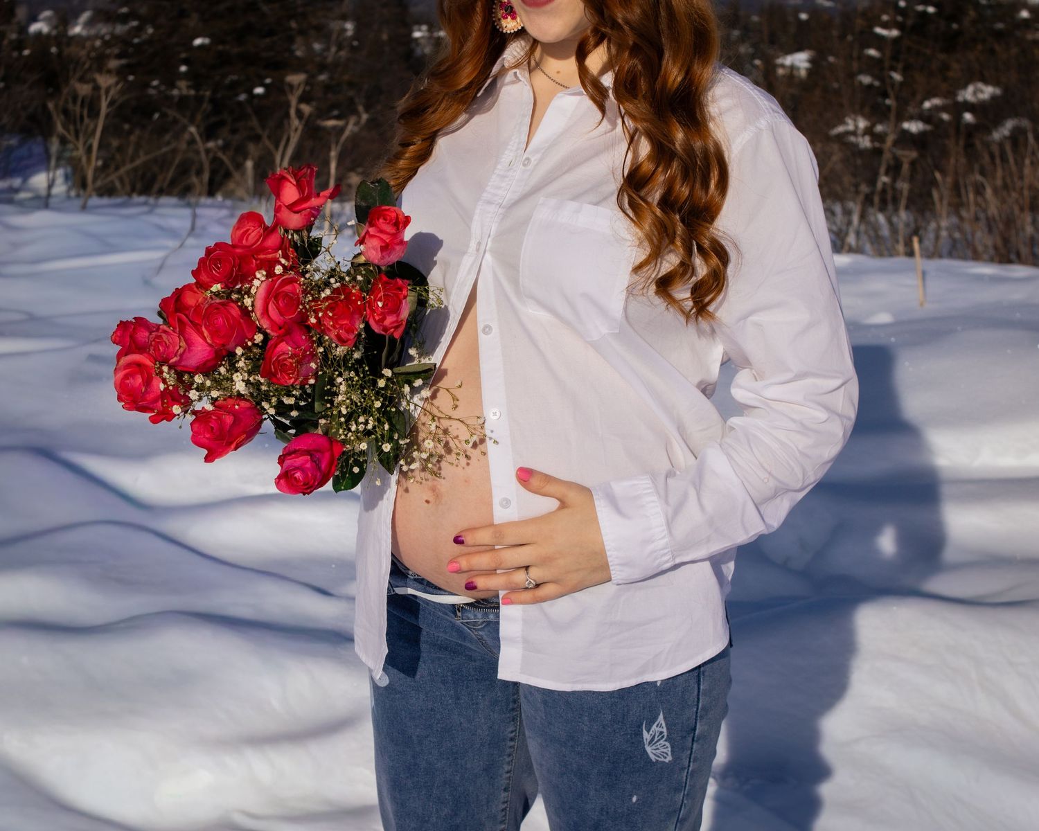 Pregnant woman in white shirt holding red roses bouquet, posing outdoors in snowy winter landscape.