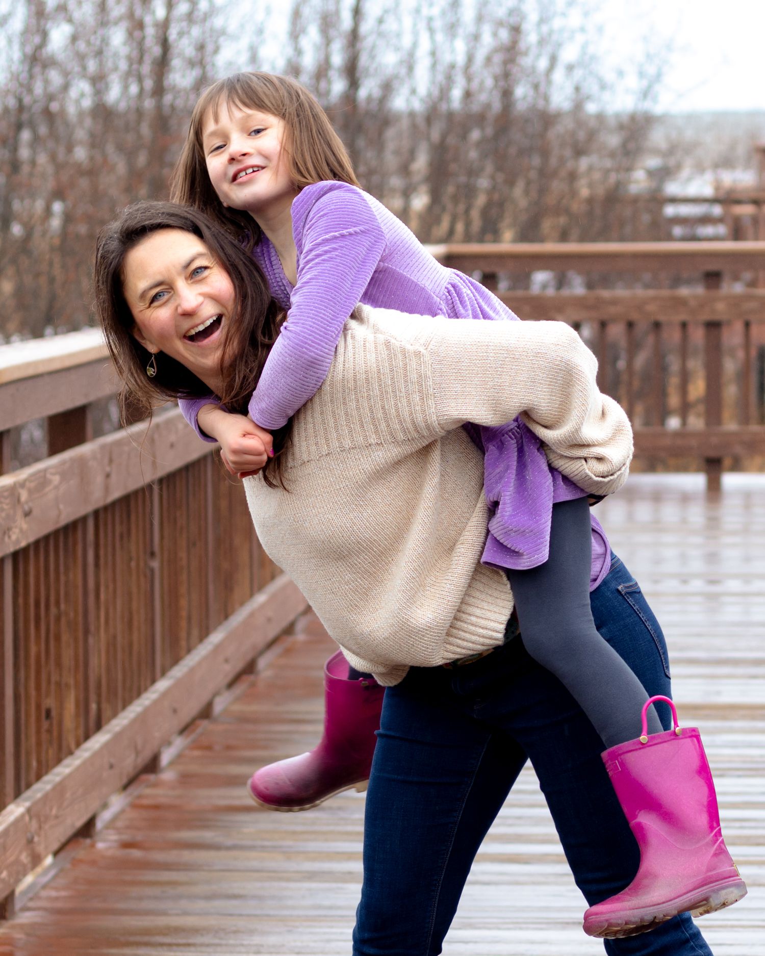 A laughing woman gives a smiling girl a piggyback ride on a wooden bridge, both wearing purple and rain boots.