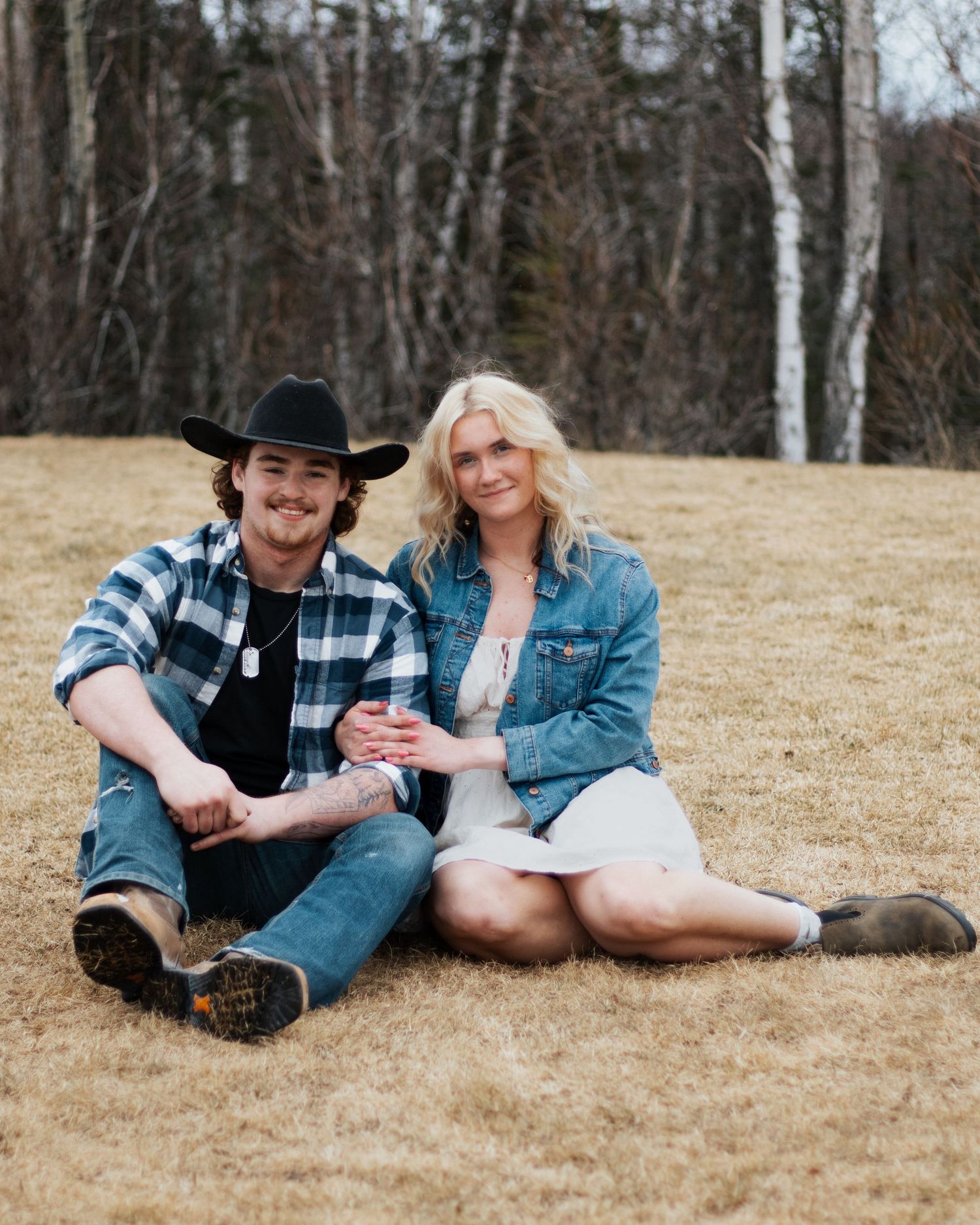 A young couple in casual western attire sits together on a grassy field, smiling, with bare trees in the background.