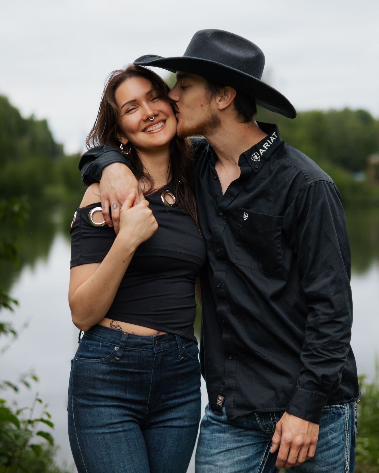 Man in black cowboy hat kissing woman's cheek outdoors near lake, both wearing black tops and jeans.