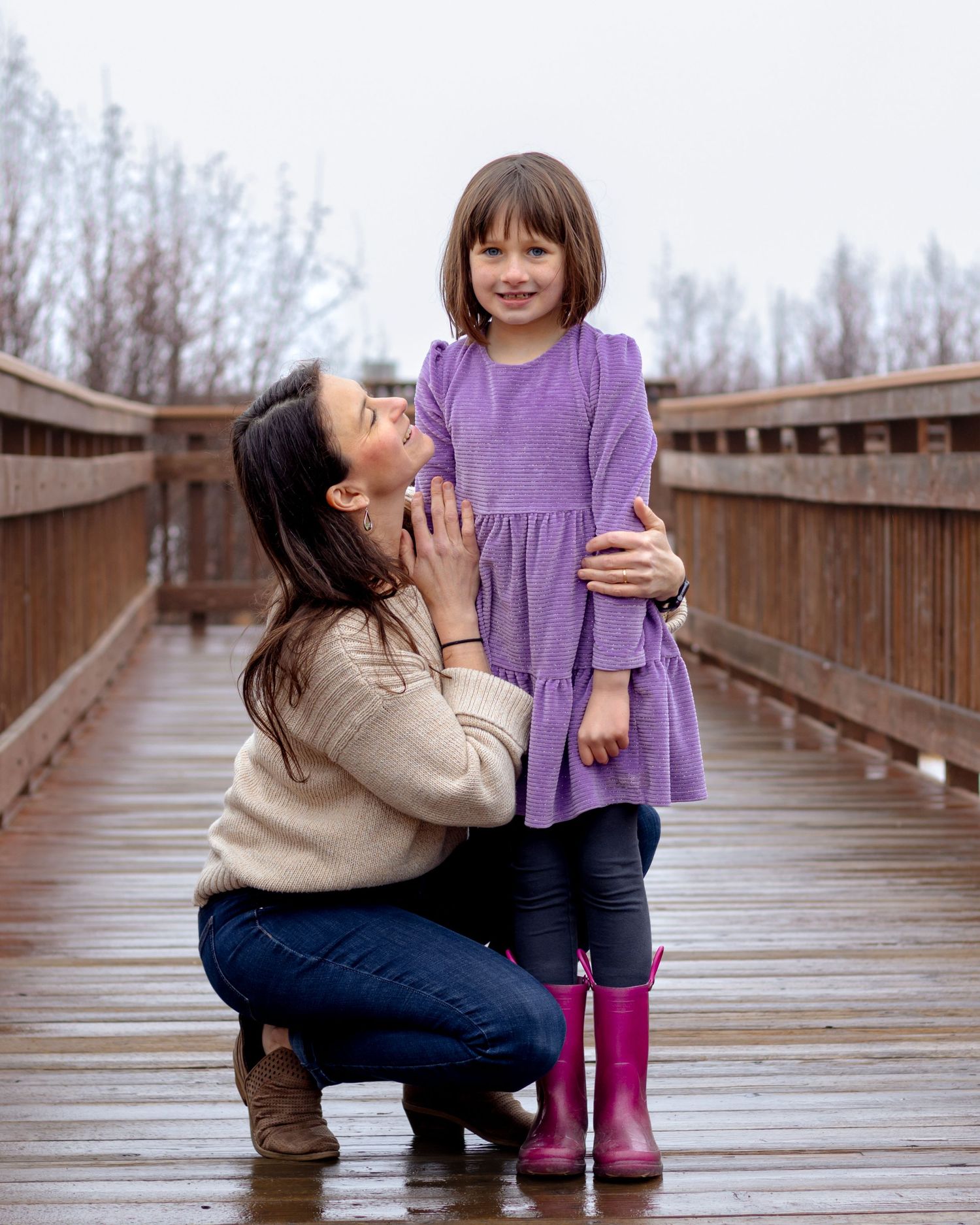A mother kneels on a wooden bridge hugging her young daughter in a purple dress and pink rain boots on a rainy day.