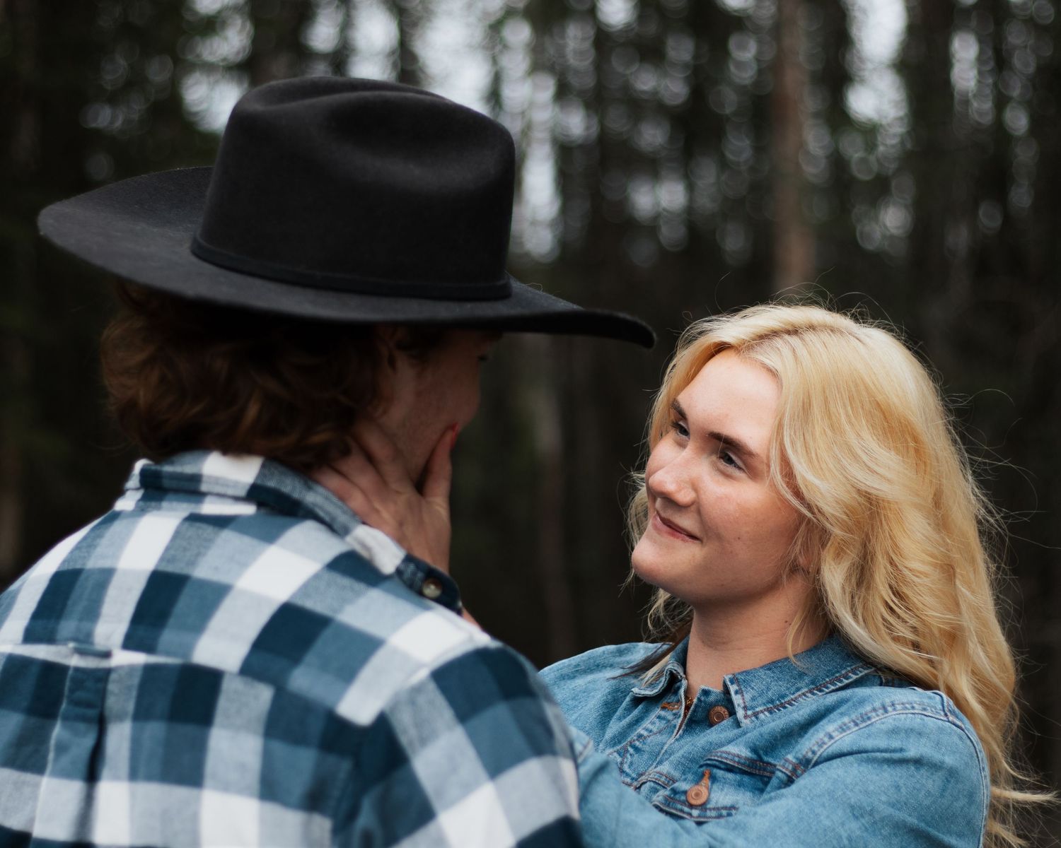 A blonde woman smiles at a man wearing a black cowboy hat and plaid shirt in a forest setting.