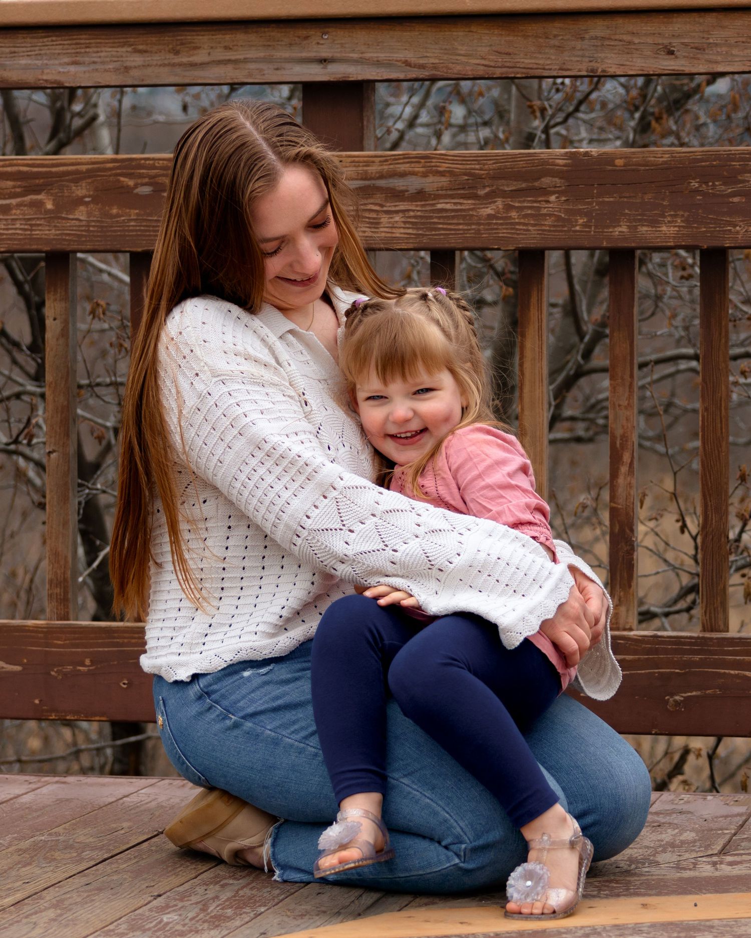 Young woman in white sweater hugging smiling toddler girl on wooden bridge outdoors.