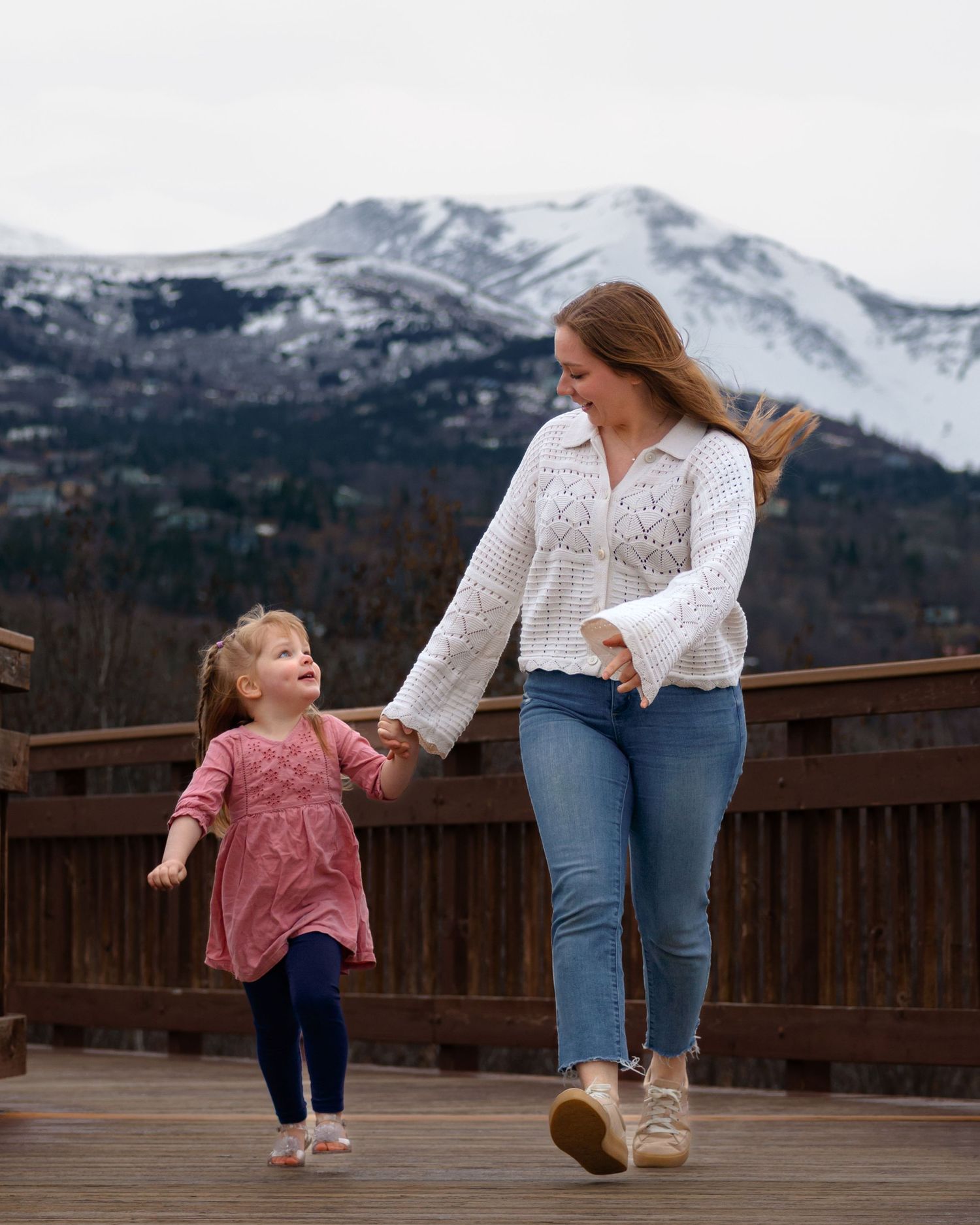 A woman and young girl hold hands walking on a wooden deck with snow-capped mountains in the background.