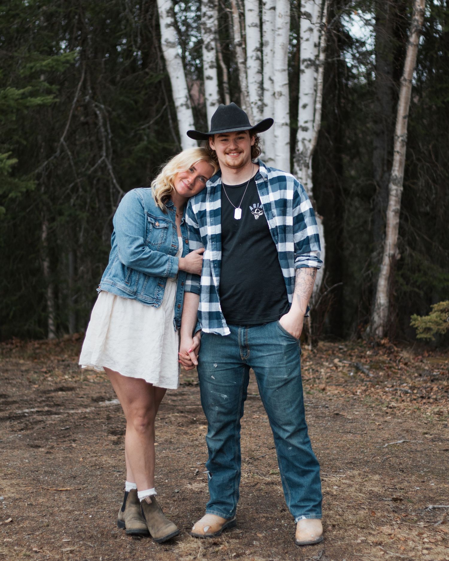 A couple poses outdoors near birch trees, woman in denim jacket and white dress, man in plaid shirt and cowboy hat.