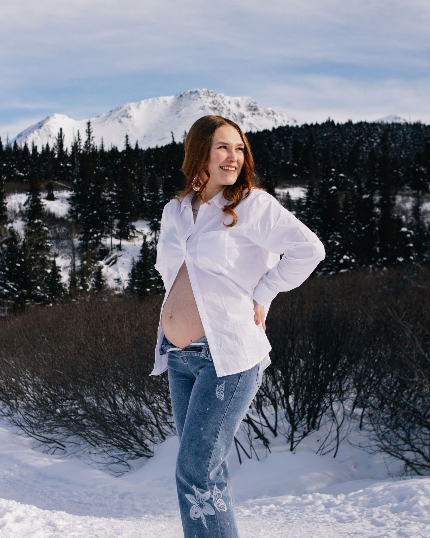 Smiling pregnant woman in white shirt and jeans posing in snowy mountain landscape with evergreen trees.