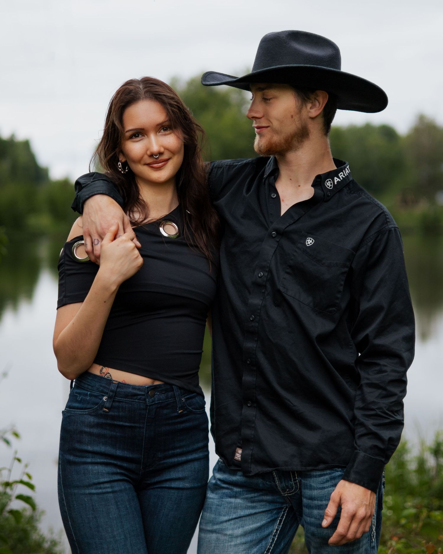 A couple in matching black outfits poses near a lake, man wearing a cowboy hat, woman in crop top and jeans.