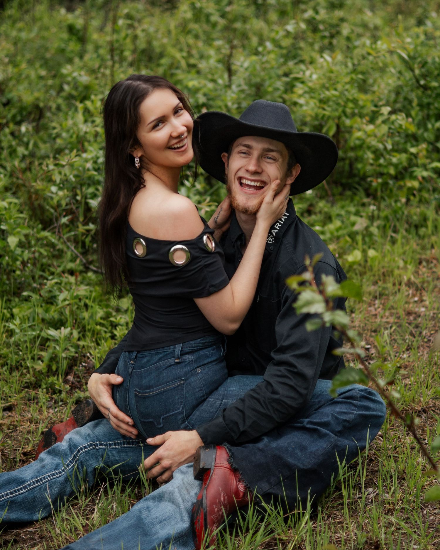 A smiling couple sits in a lush green field, woman in black off-shoulder top embraces man in black cowboy hat.