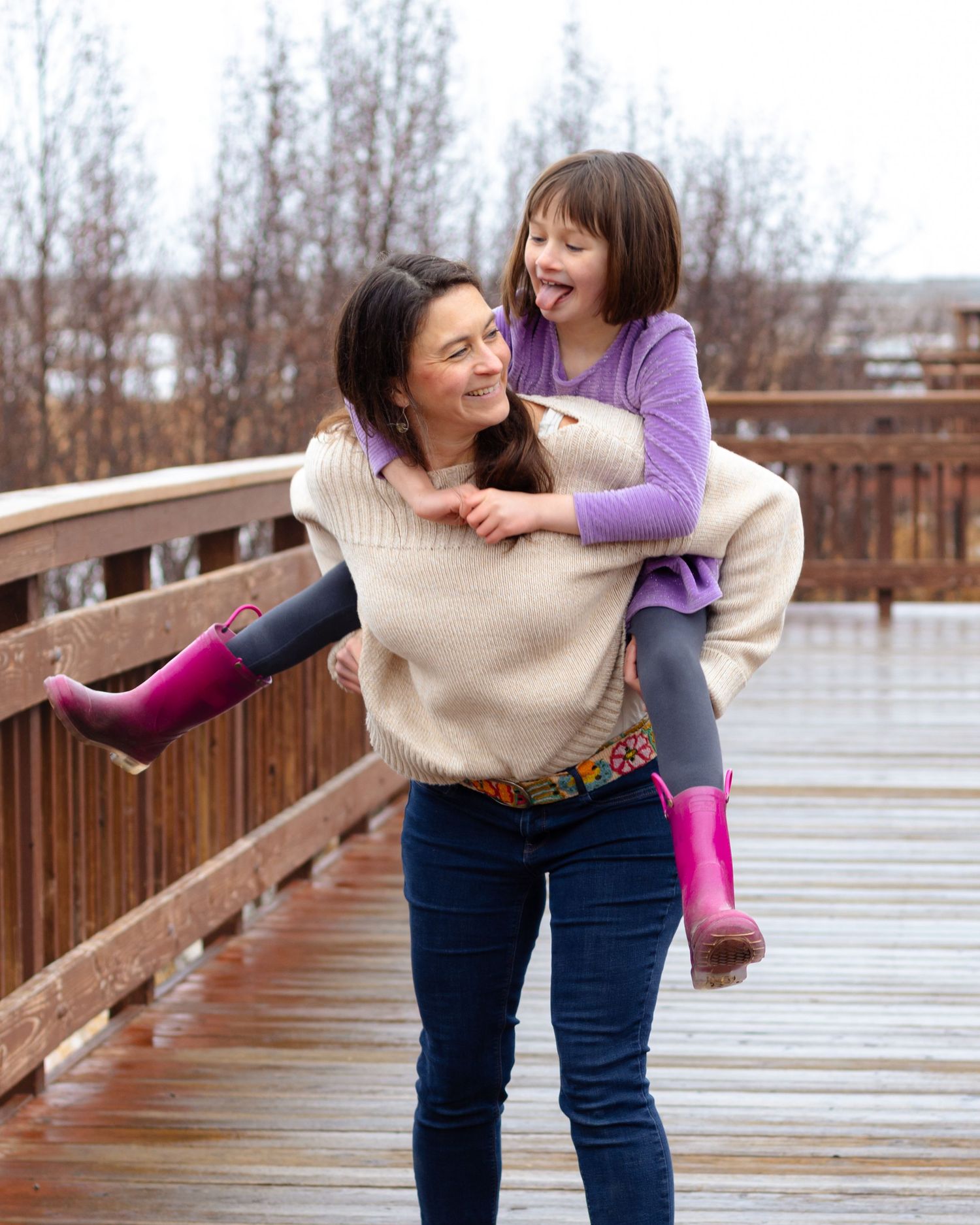 Woman giving laughing girl a piggyback ride on wooden bridge, both wearing purple boots in rainy weather.