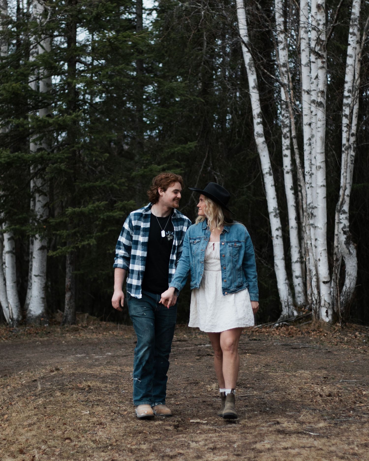 Couple holding hands and smiling at each other while walking on a forest path surrounded by birch trees.