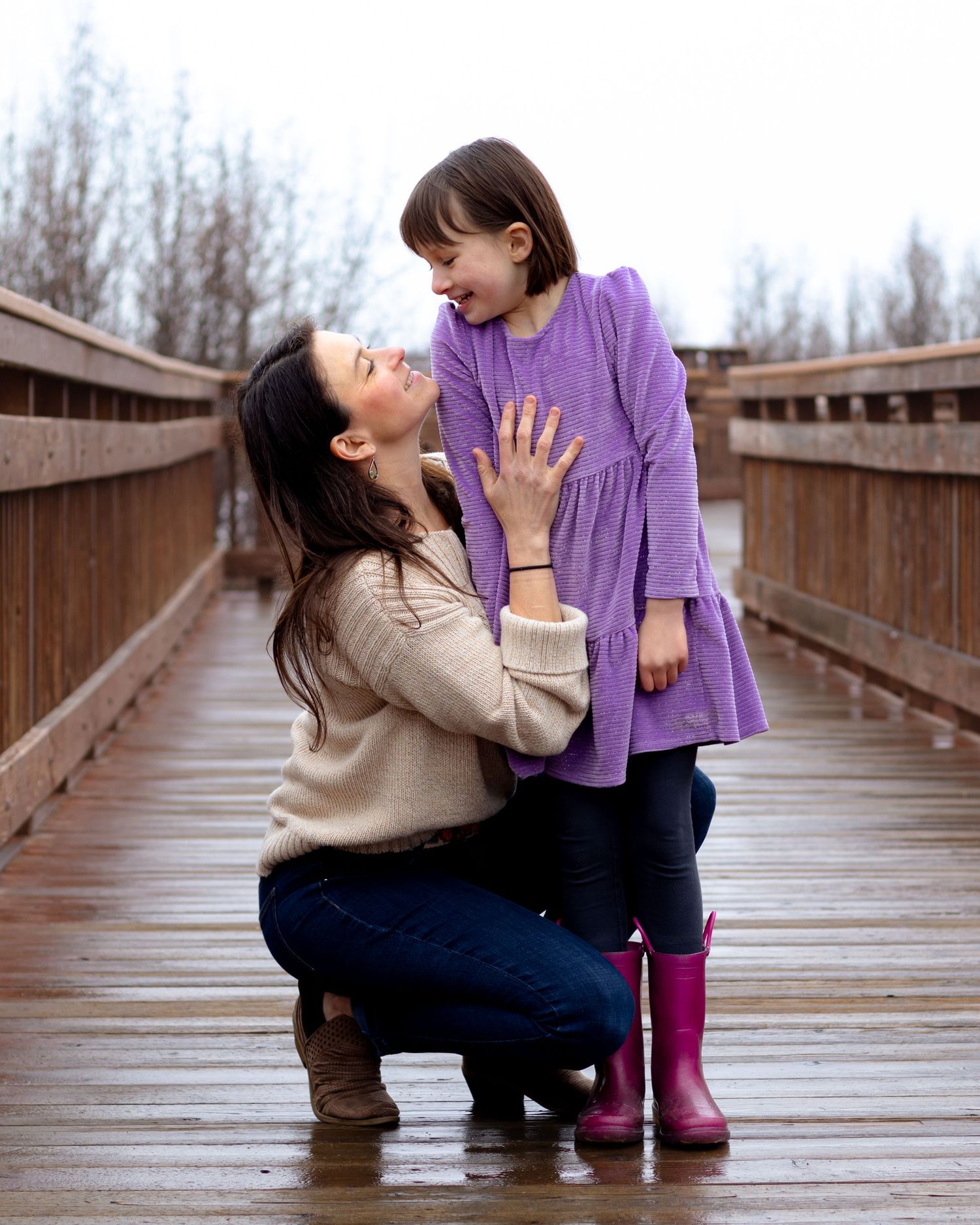 A mother kneels on a wooden bridge smiling up at her laughing young daughter in a purple dress and pink boots.