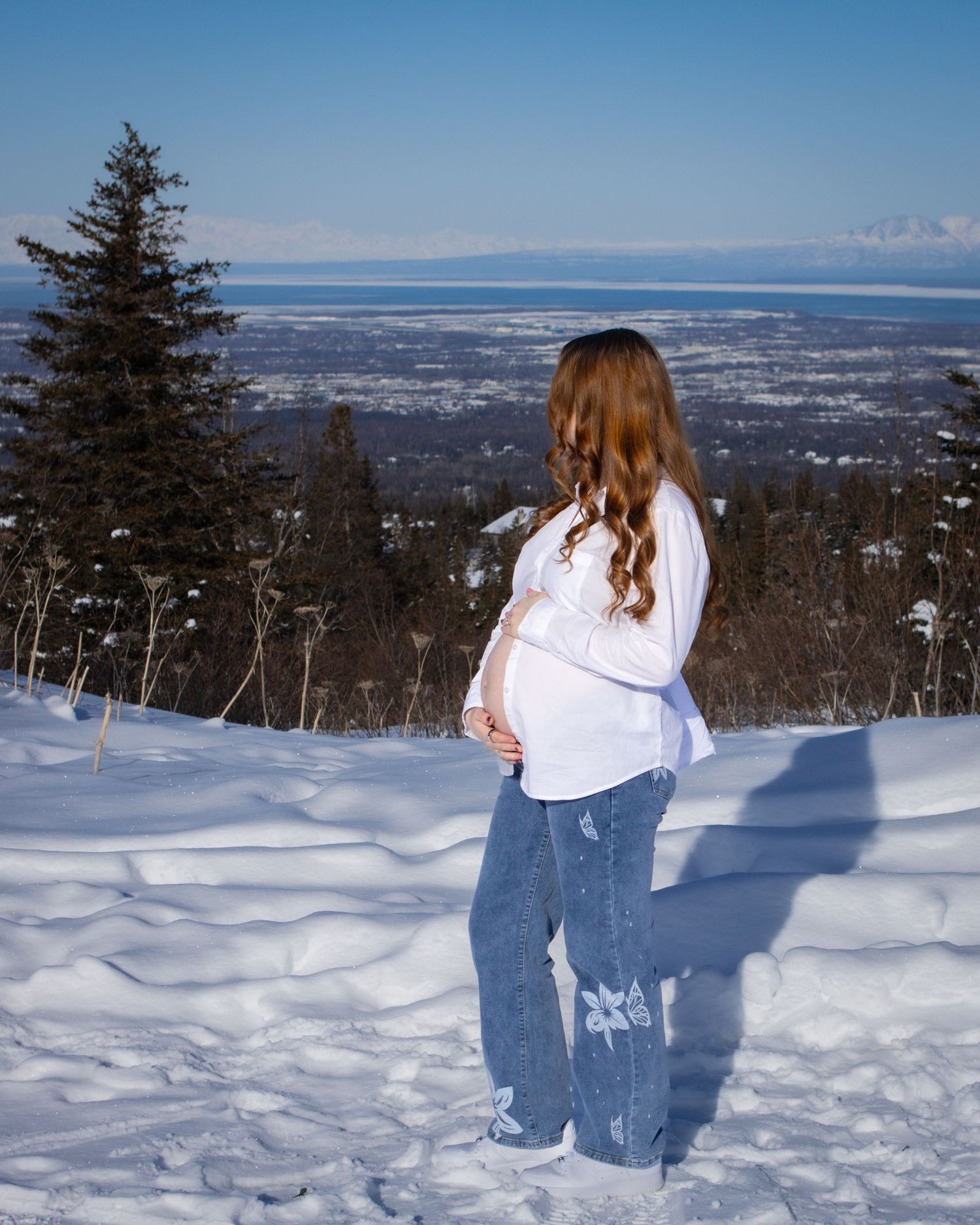 Pregnant woman in white top and jeans stands on snowy hillside overlooking a coastal city on a clear winter day.