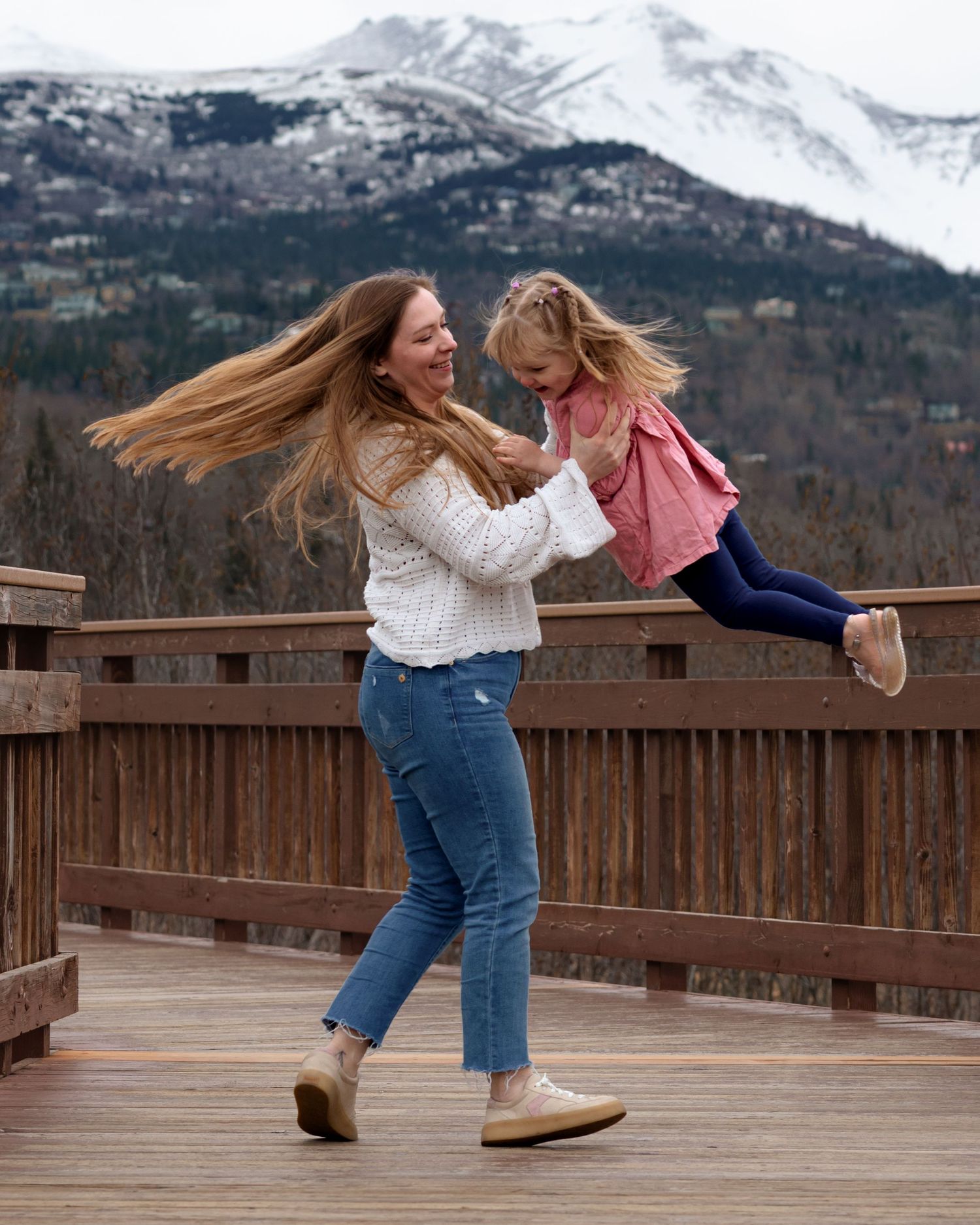 A woman with flowing hair joyfully lifts a young girl on a wooden deck with snowy mountains in the background.