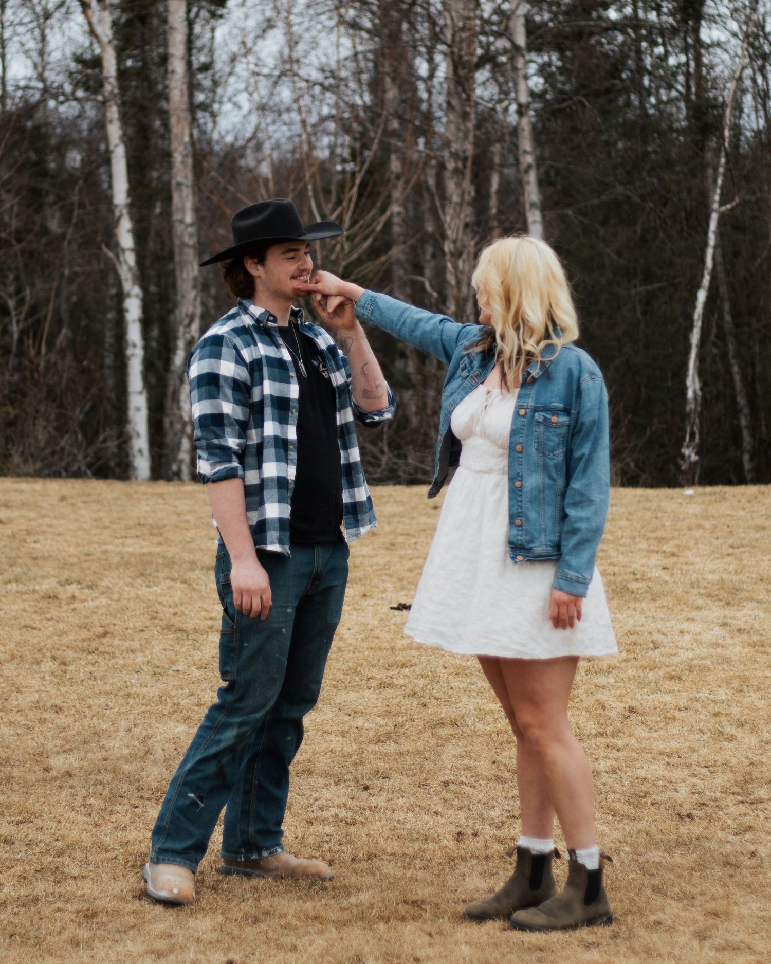 A couple in casual western attire shares a playful moment in a rustic field with bare trees in the background.