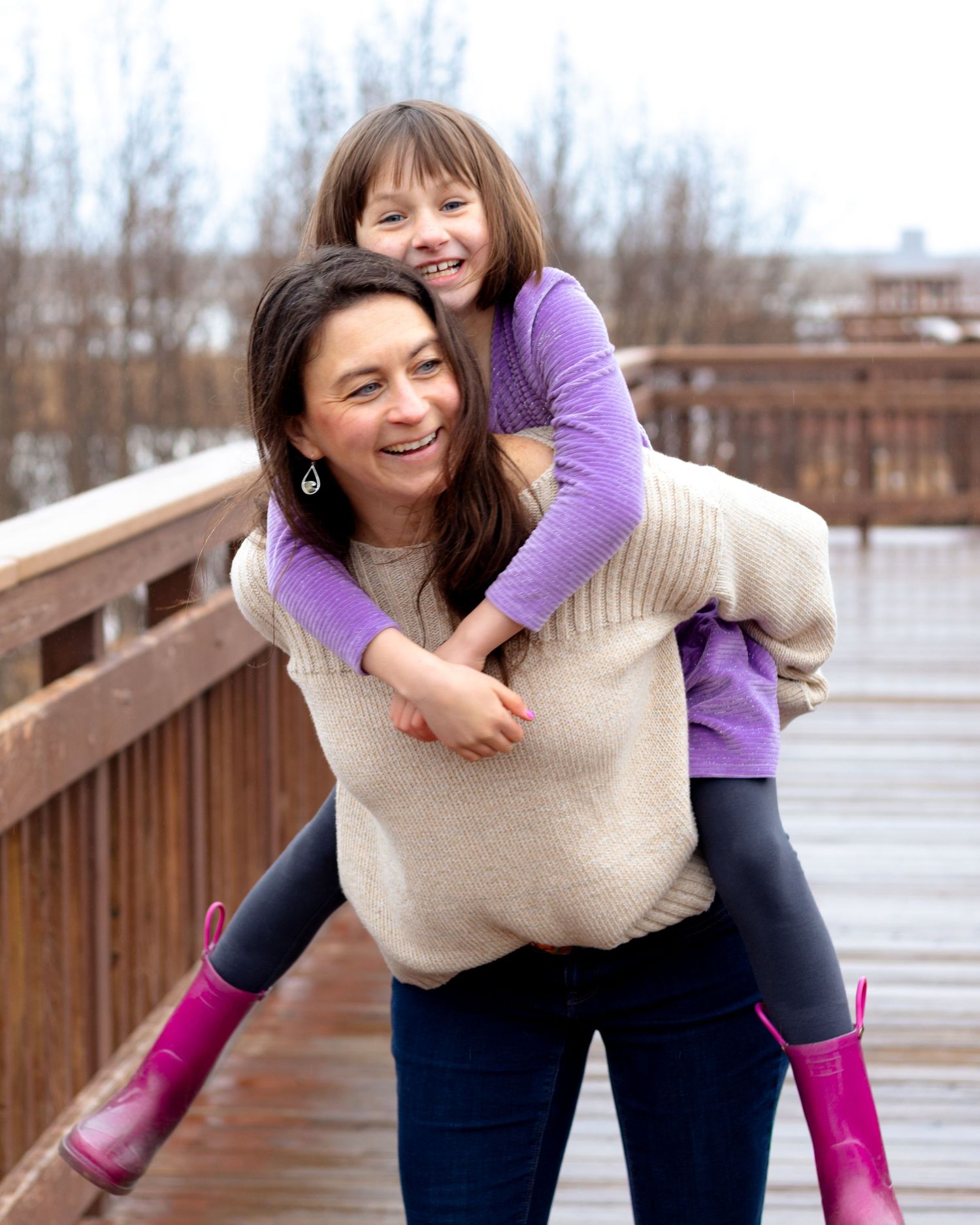 A woman gives a smiling girl a piggyback ride on a wooden bridge, both wearing purple and pink rain boots.