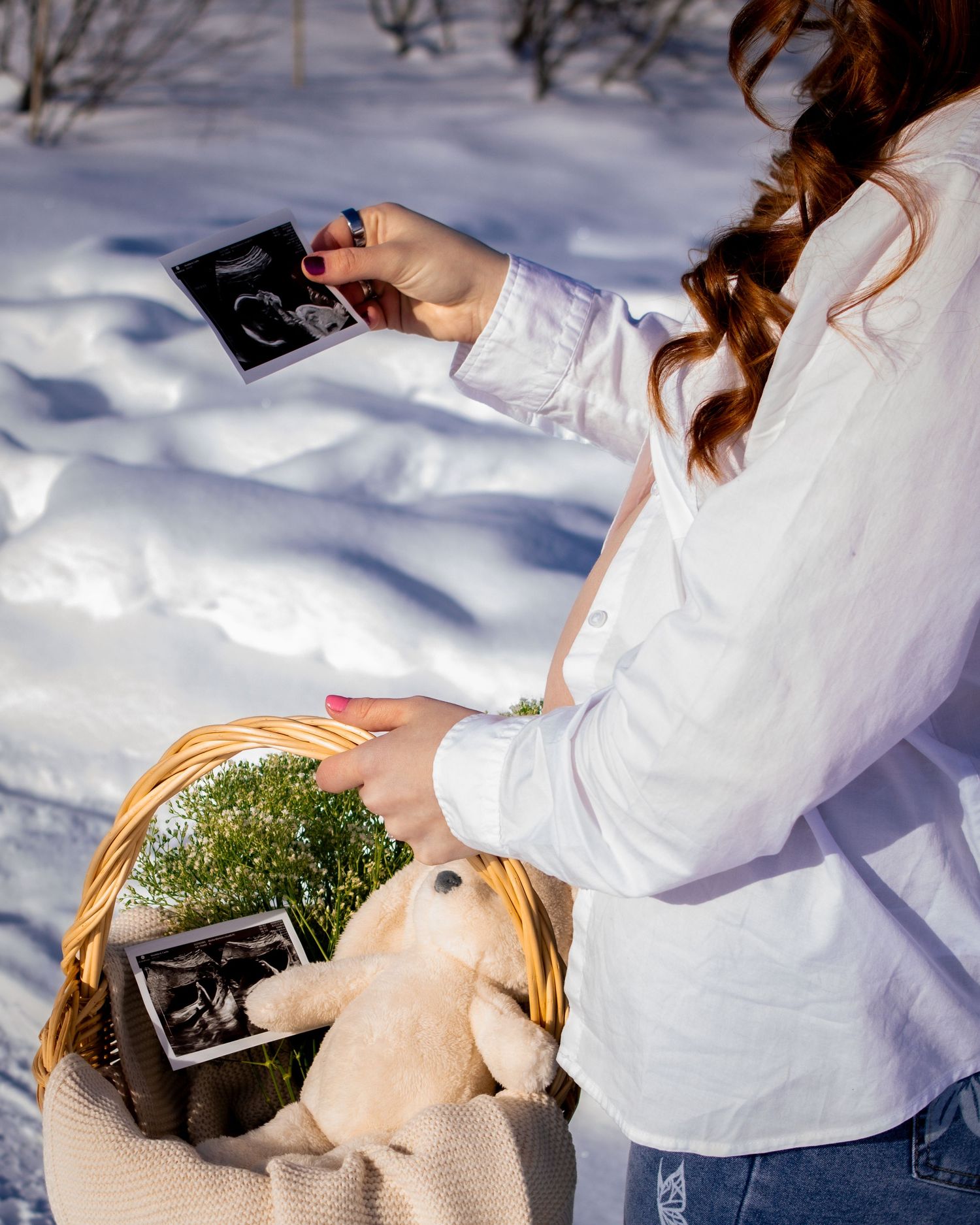 Pregnant woman in white shirt holds ultrasound photos over basket with stuffed bunny in snowy winter setting.