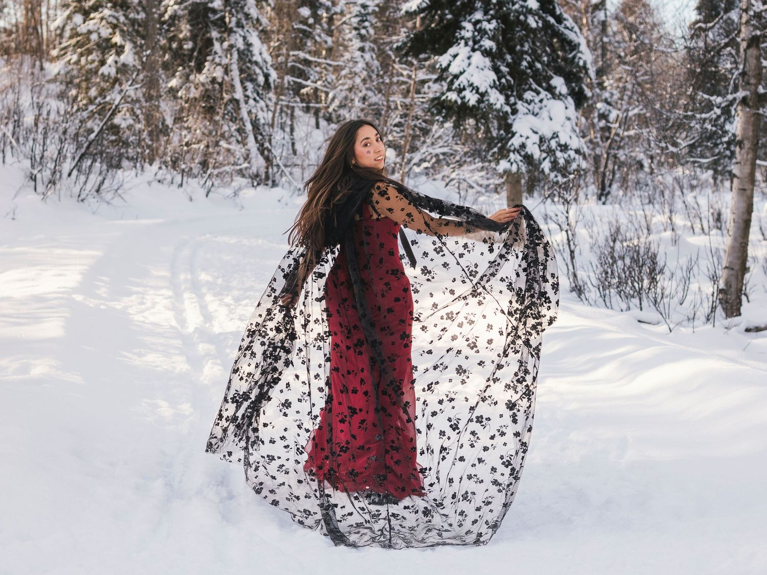 Woman in red dress with black lace shawl posing gracefully in snowy winter forest landscape.