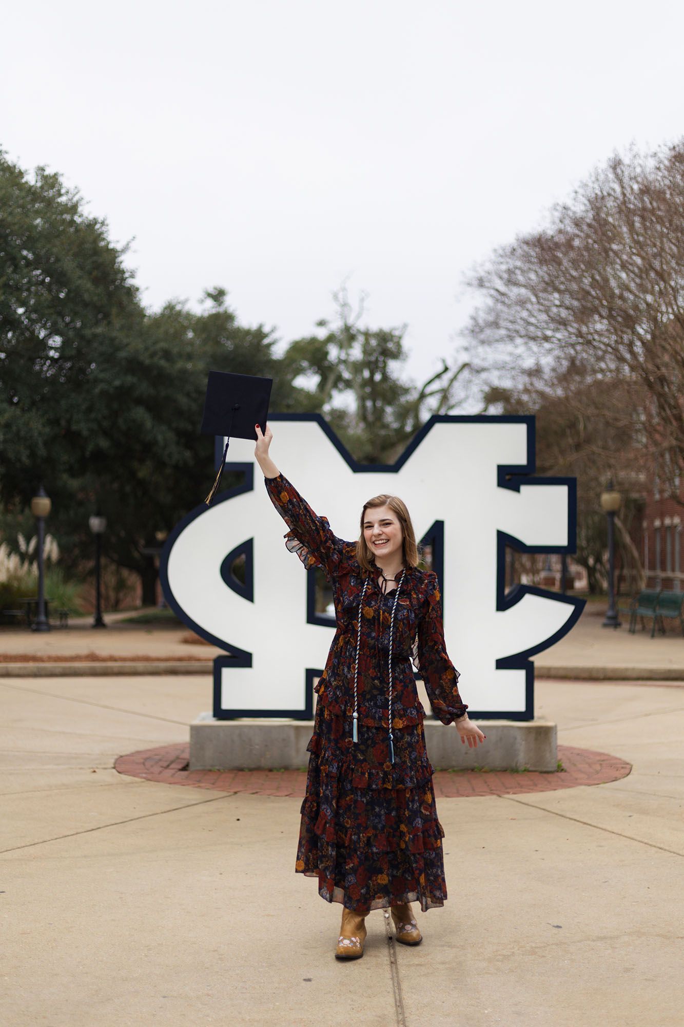 Female graduate celebrates in front of MC university logo sign, holding cap aloft in floral maxi dress.