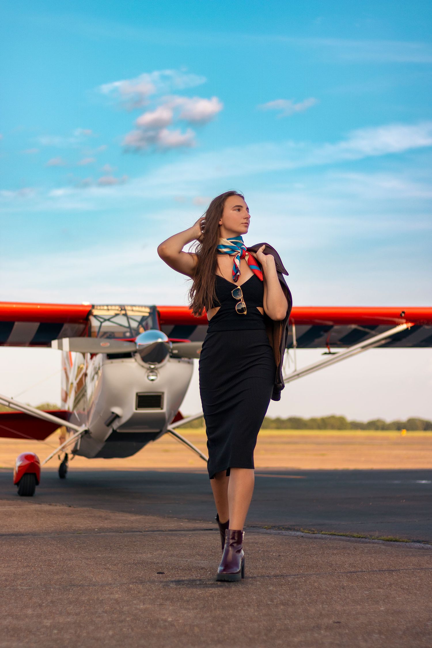 Elegant woman in black dress with colorful scarf walking on airport tarmac near small red plane.