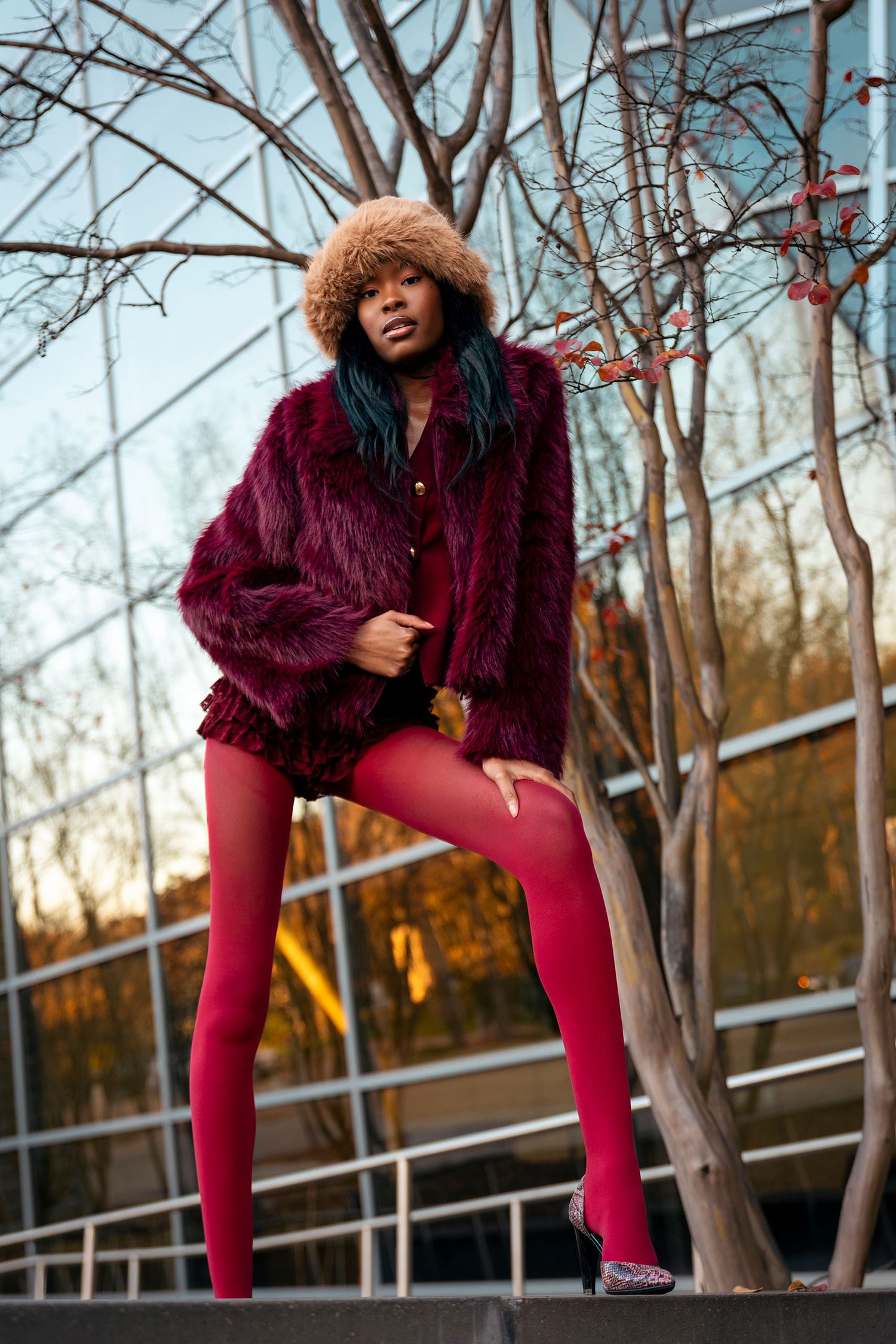 Woman in burgundy fur coat and red tights poses near bare tree at sunset.