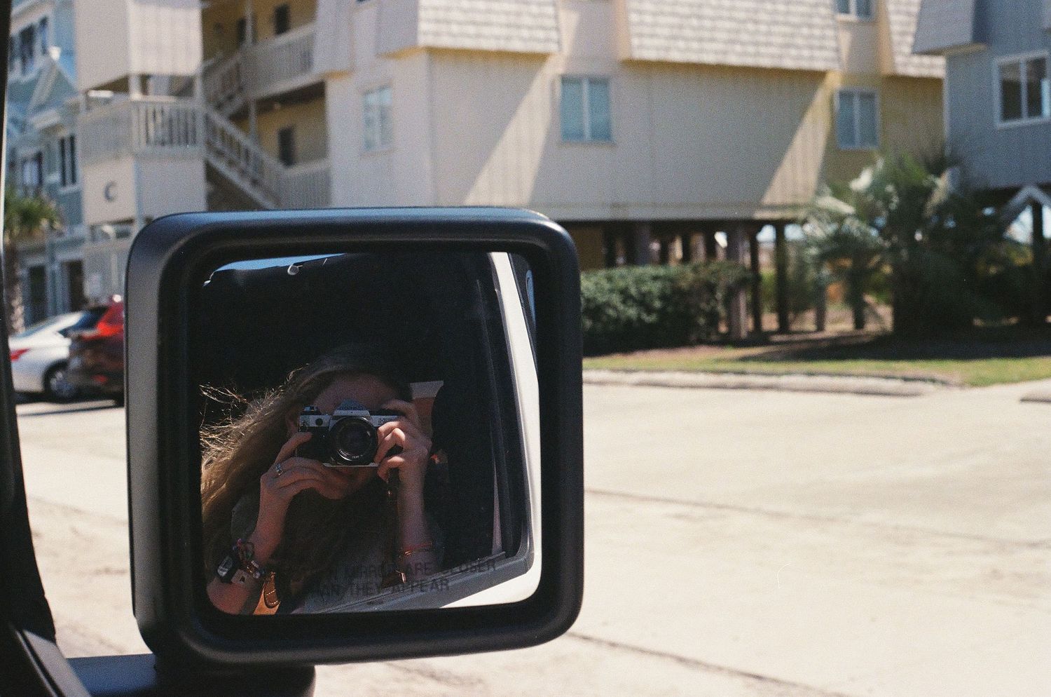 A woman photographs herself in a vehicle side mirror on a sunny suburban street.
