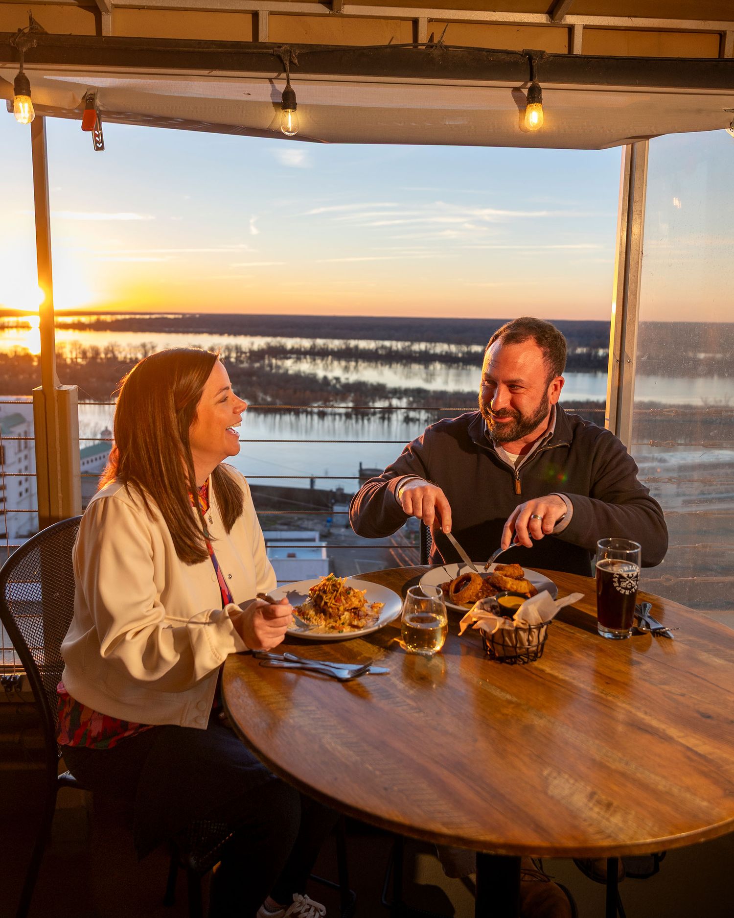 Couple dining at a rooftop restaurant enjoying food and drinks with a stunning sunset river view.