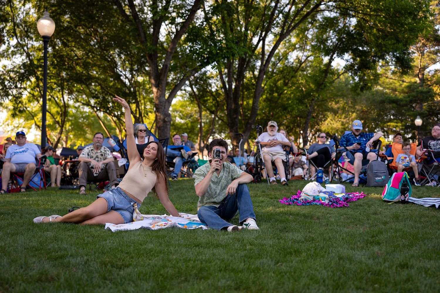 Couple sits on grass enjoying an outdoor event in a shaded park with a crowd behind them.