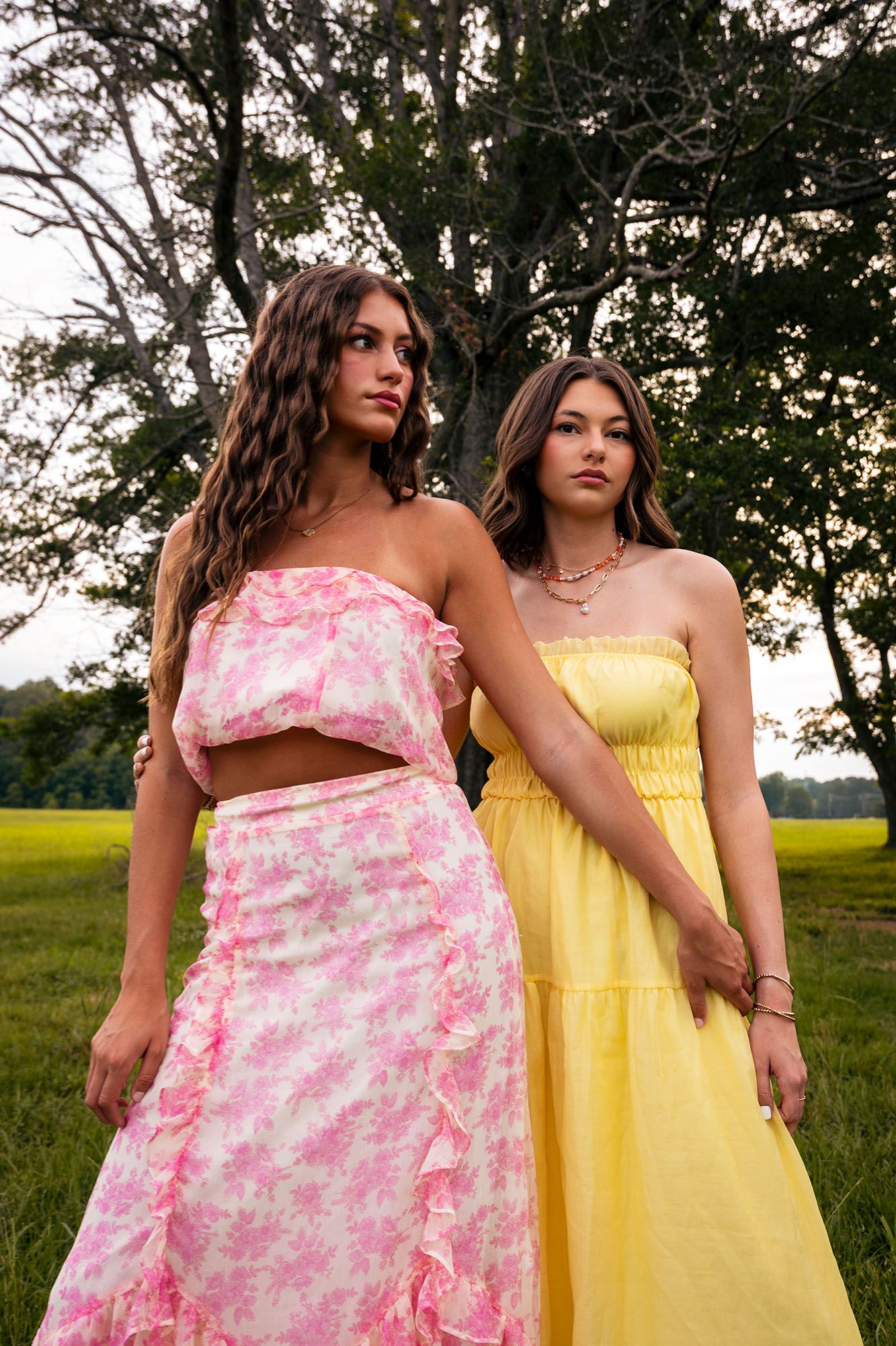 Two young women in pink floral and yellow sundresses pose in green meadow by trees.