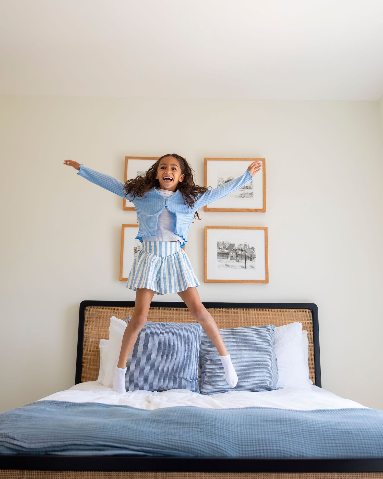 A joyful young girl jumps on a bed wearing a blue cardigan and striped skirt in a bright bedroom.