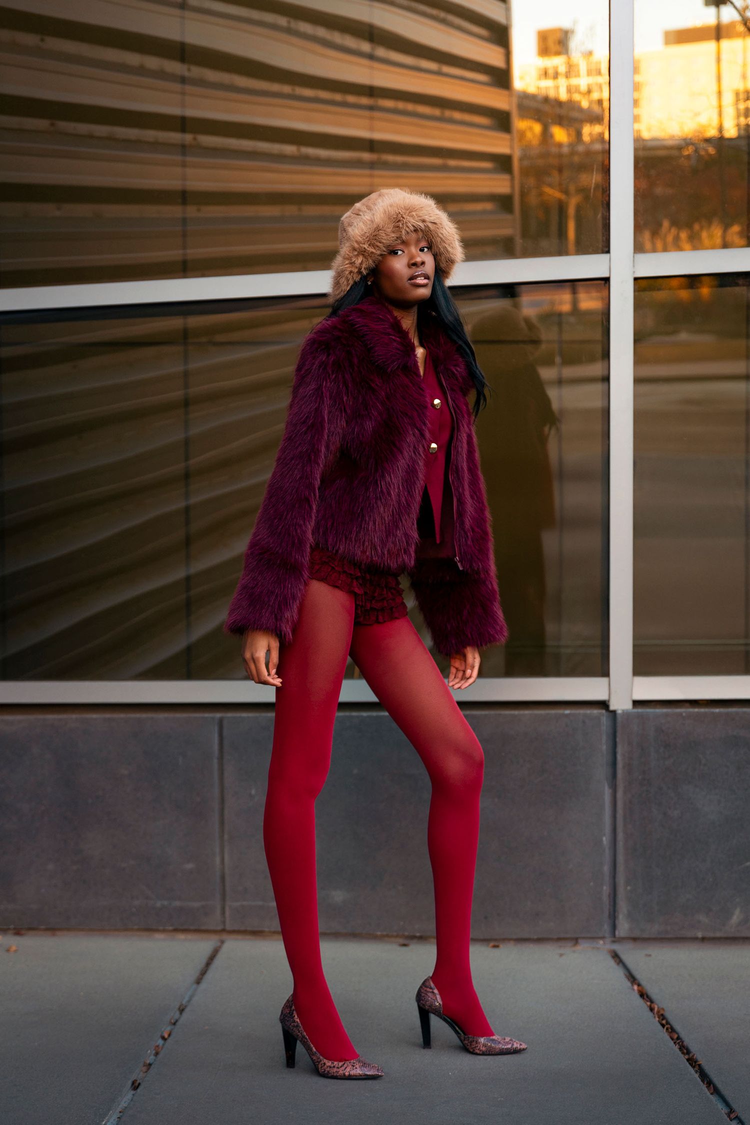 Woman in purple fur coat and red tights poses against glass building exterior.