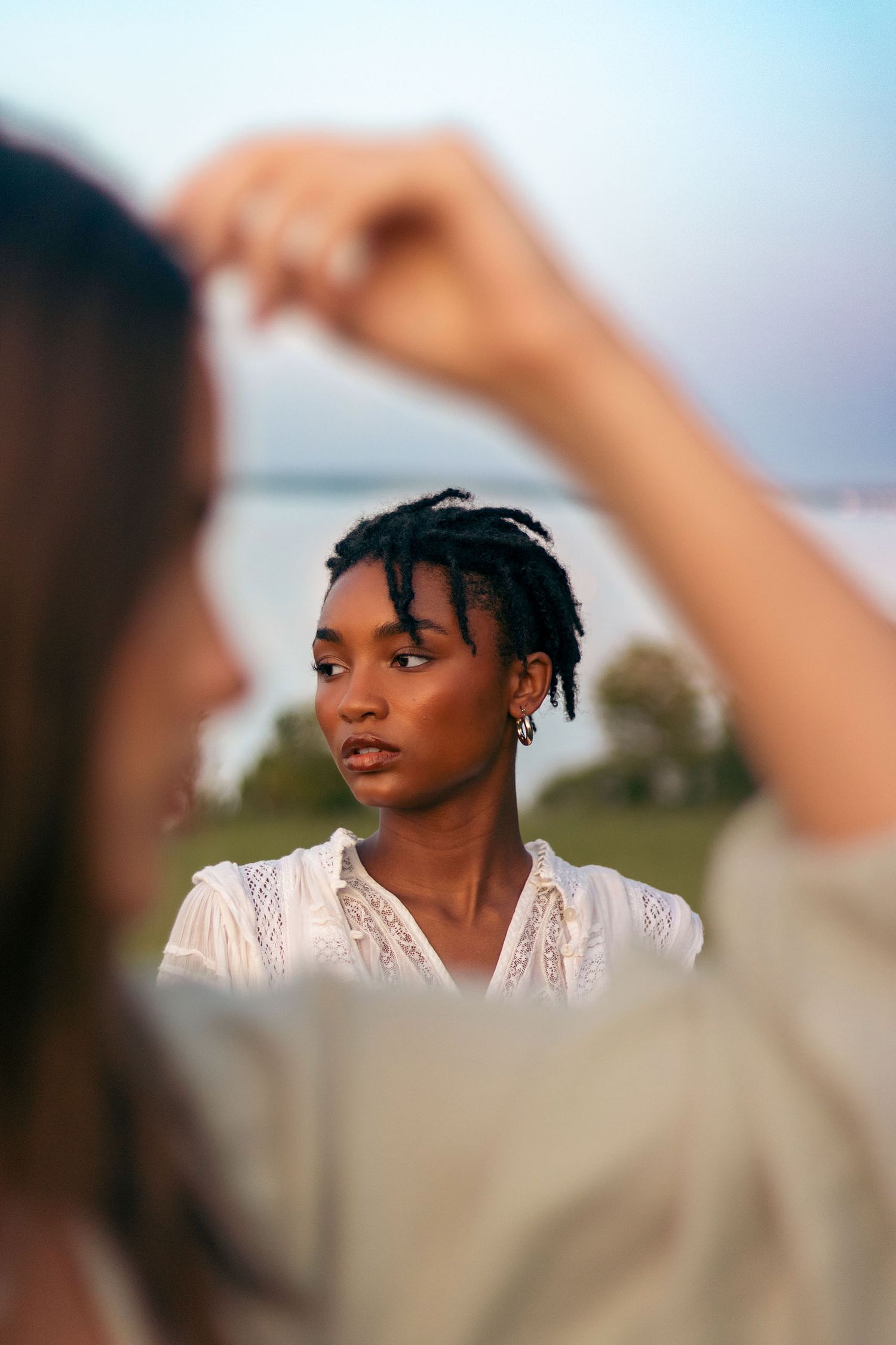 Young Black woman with short dreadlocks in white blouse photographed through blurred foreground outdoors.