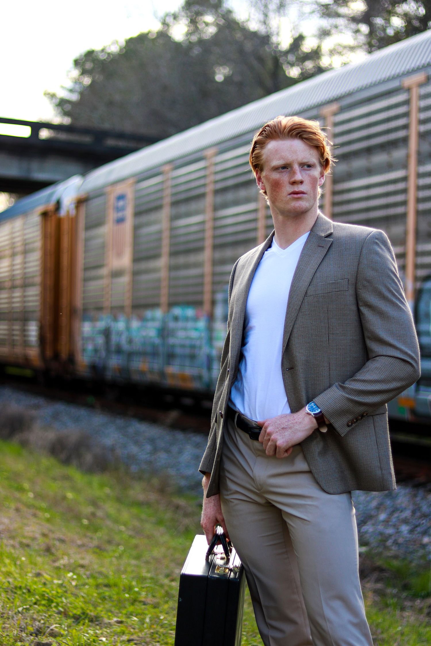 Stylish redhead man in gray suit holding briefcase standing near graffiti freight train outdoors.