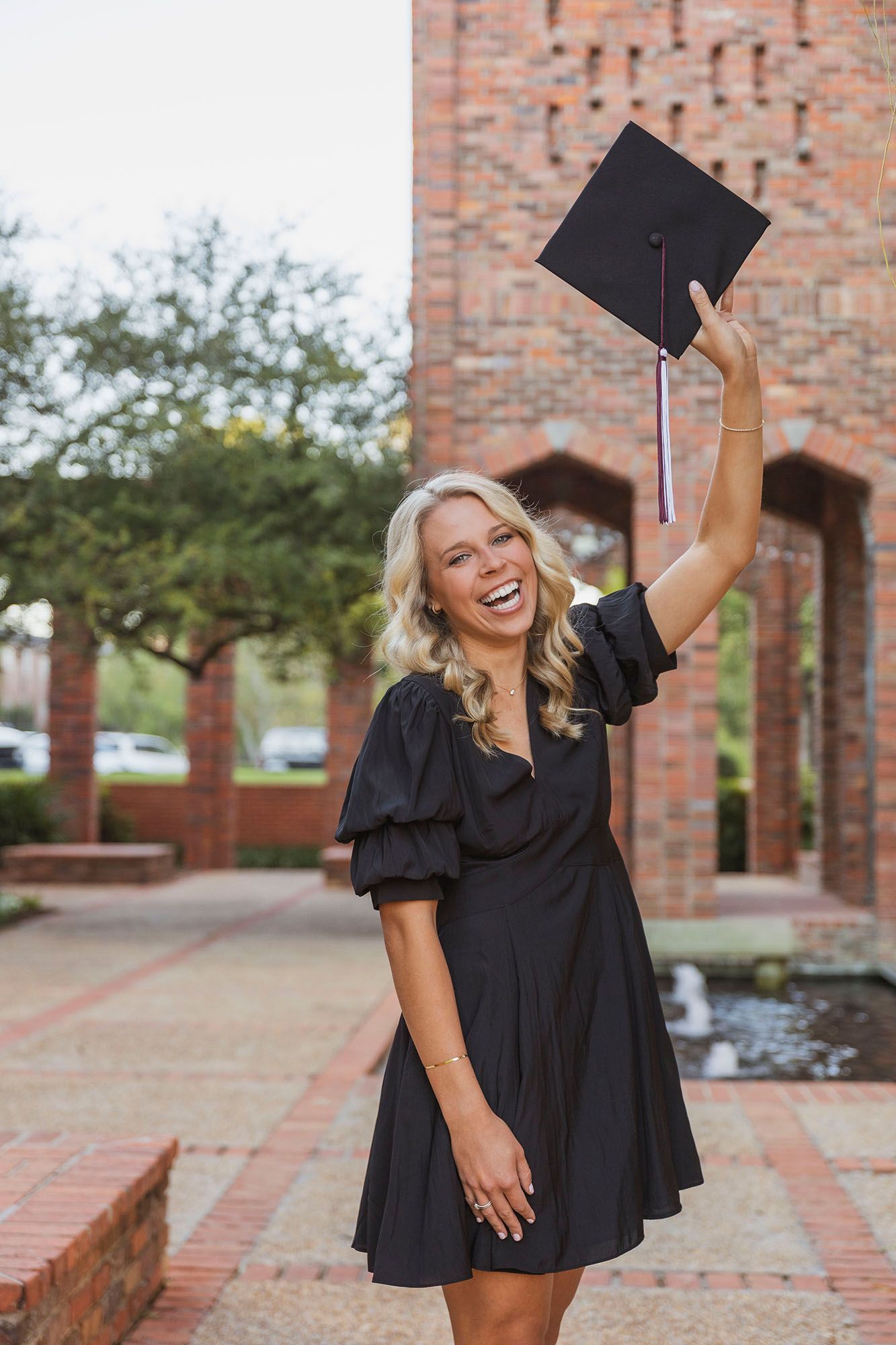 Smiling blonde female graduate in black dress raises cap triumphantly near brick university building.