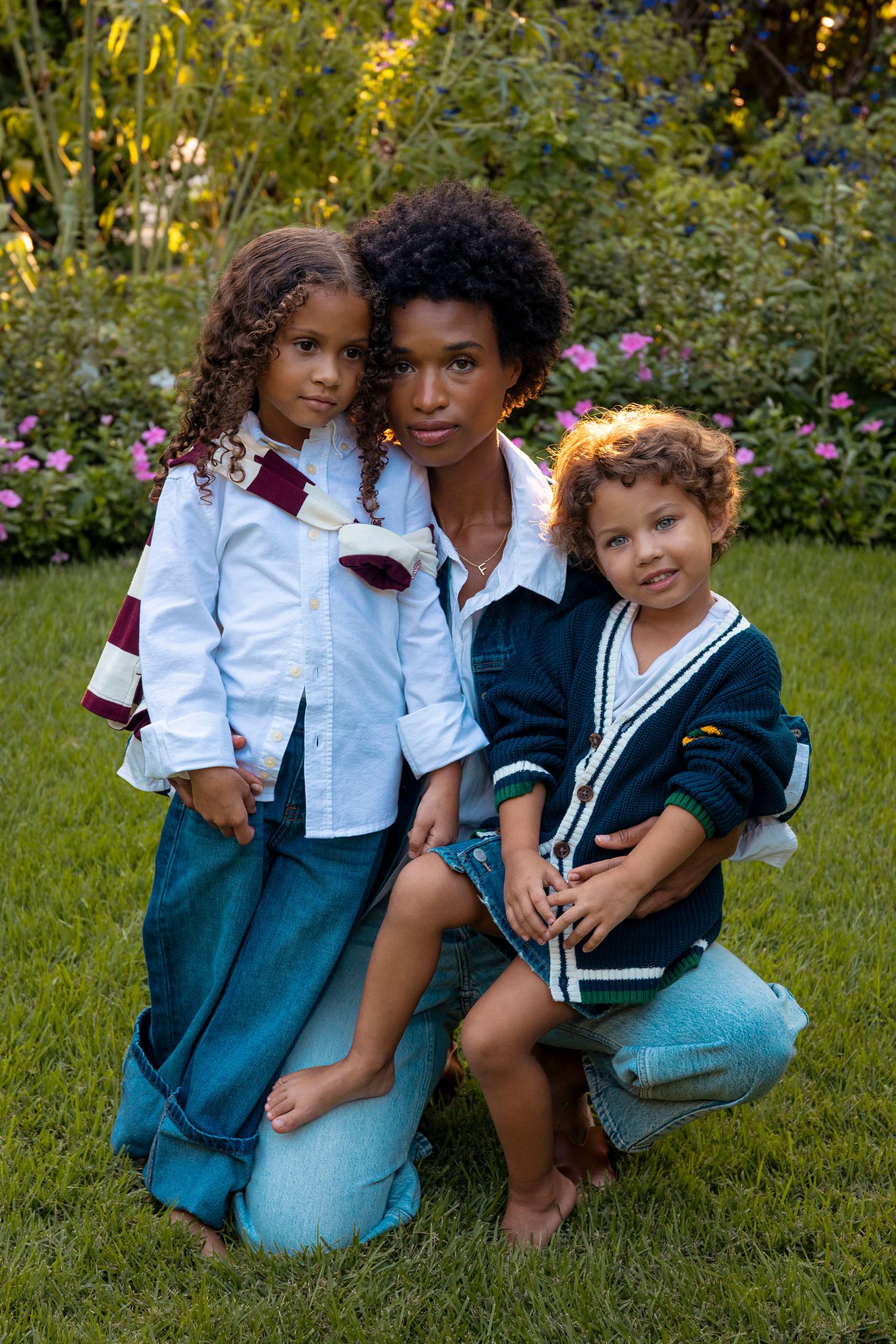 A mother with natural curly hair poses with her two young children in a lush garden setting.