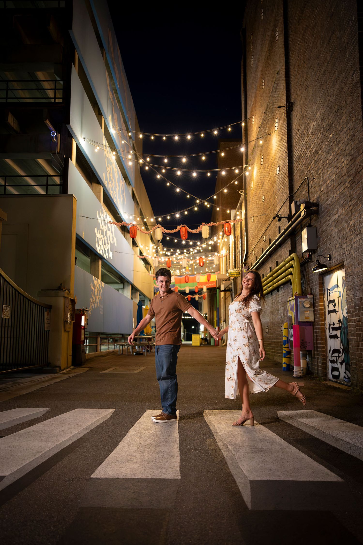 Couple holds hands crossing a striped crosswalk in a festive alley lit with string lights at night.
