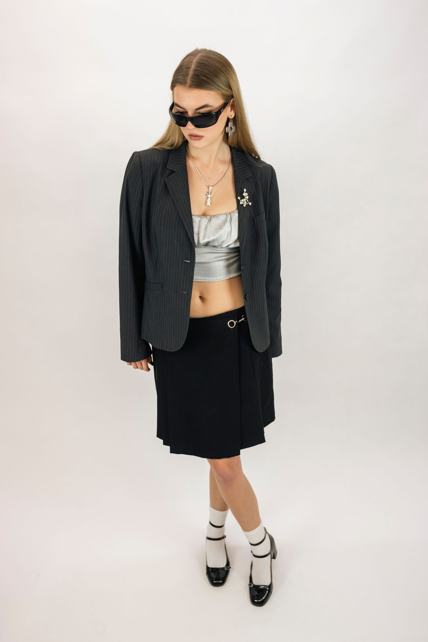Woman in black blazer, skirt, silver crop top, sunglasses, and white socks with Mary Jane heels poses against white background.